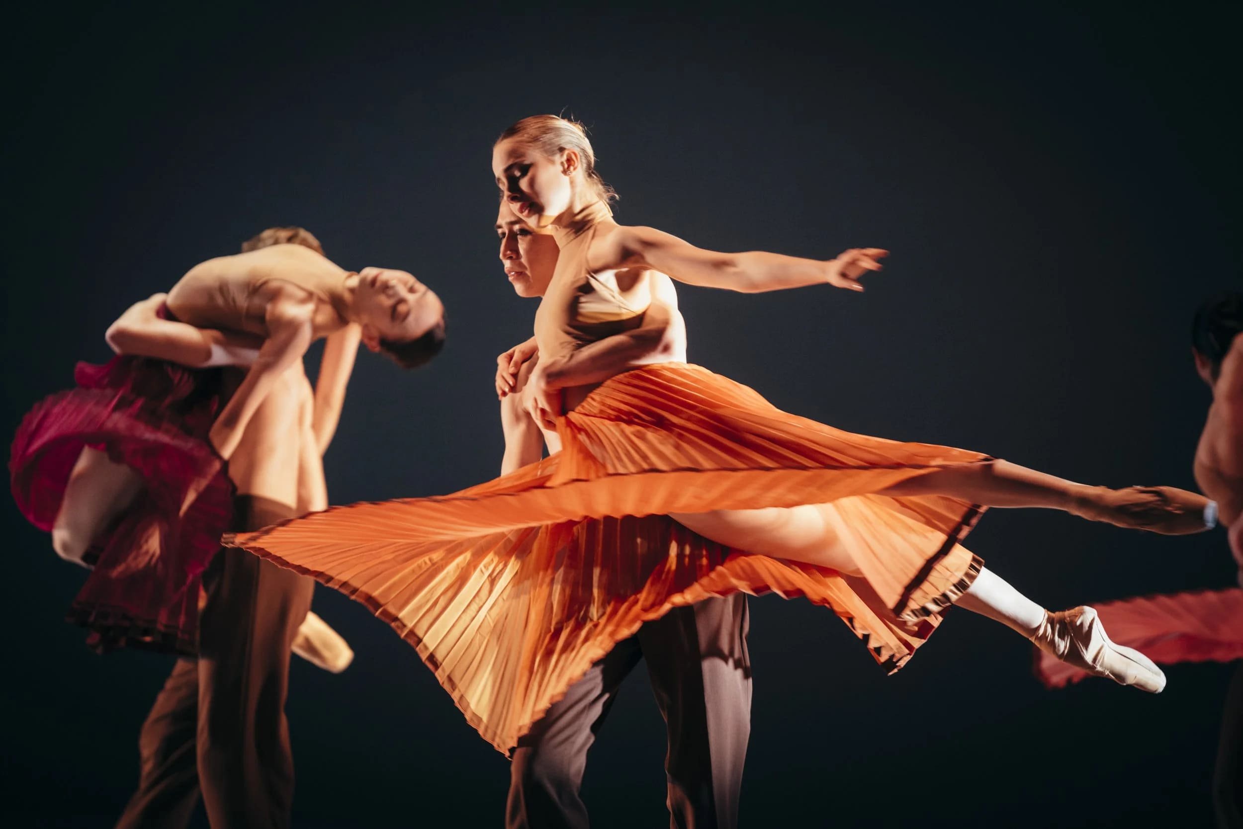 Dancer in an orange pleated skirt extends a leg while being supported by a partner on stage under warm theatre lighting.