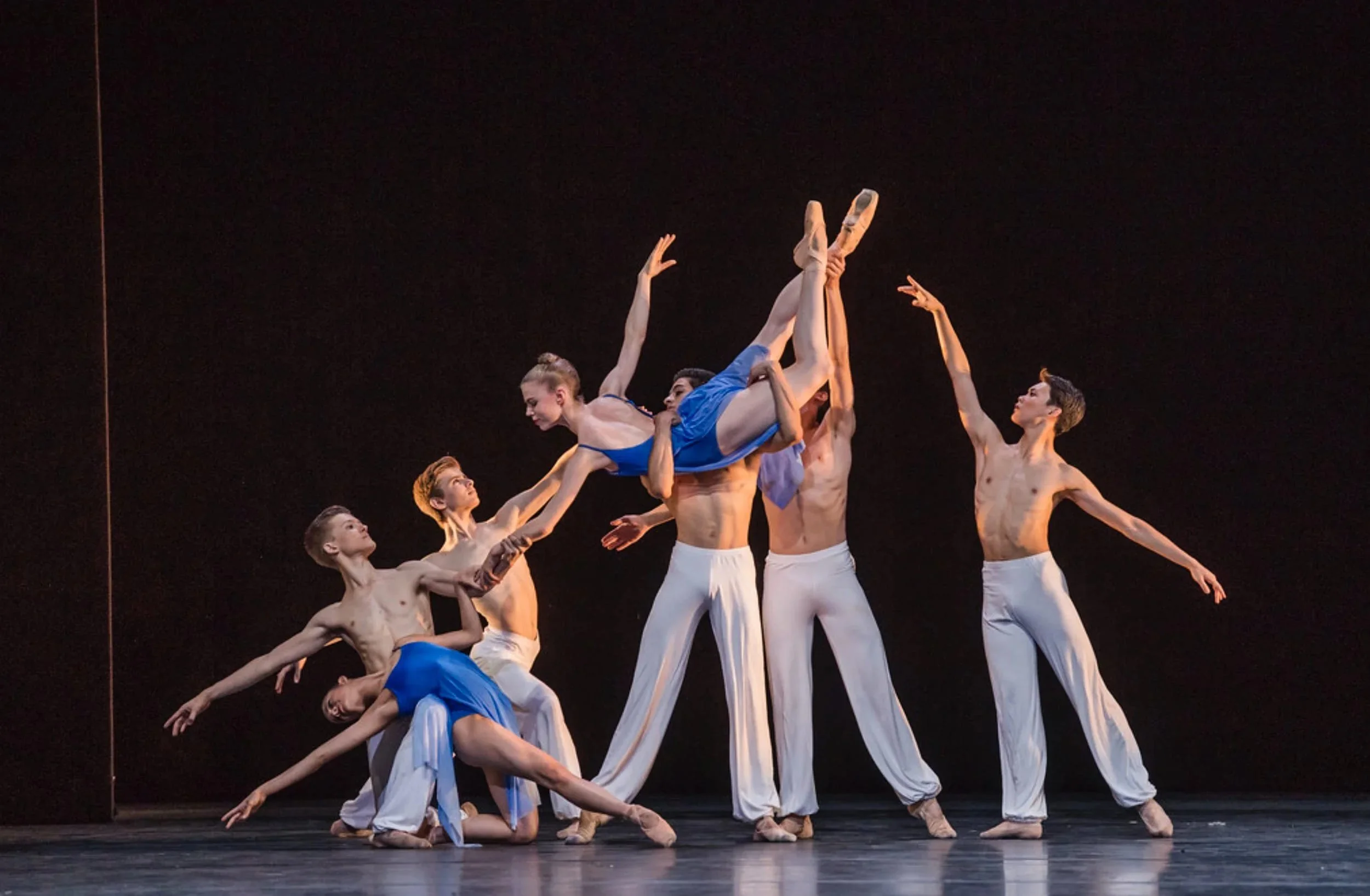 Ballet ensemble on stage with dancers in blue costumes and white trousers; one dancer is lifted horizontally above the group.