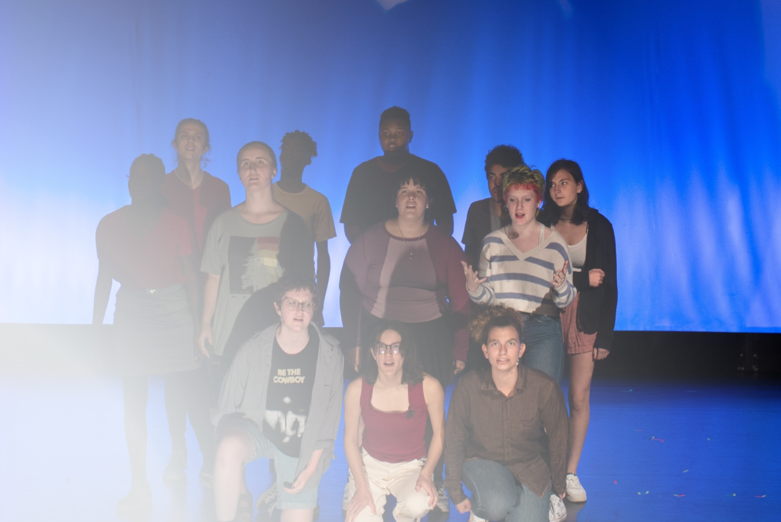 A group of young people dressed in street clothes on stage ,  with some standing and others in a crouched position, under blue lighting. | Yes! And Collaborative Arts