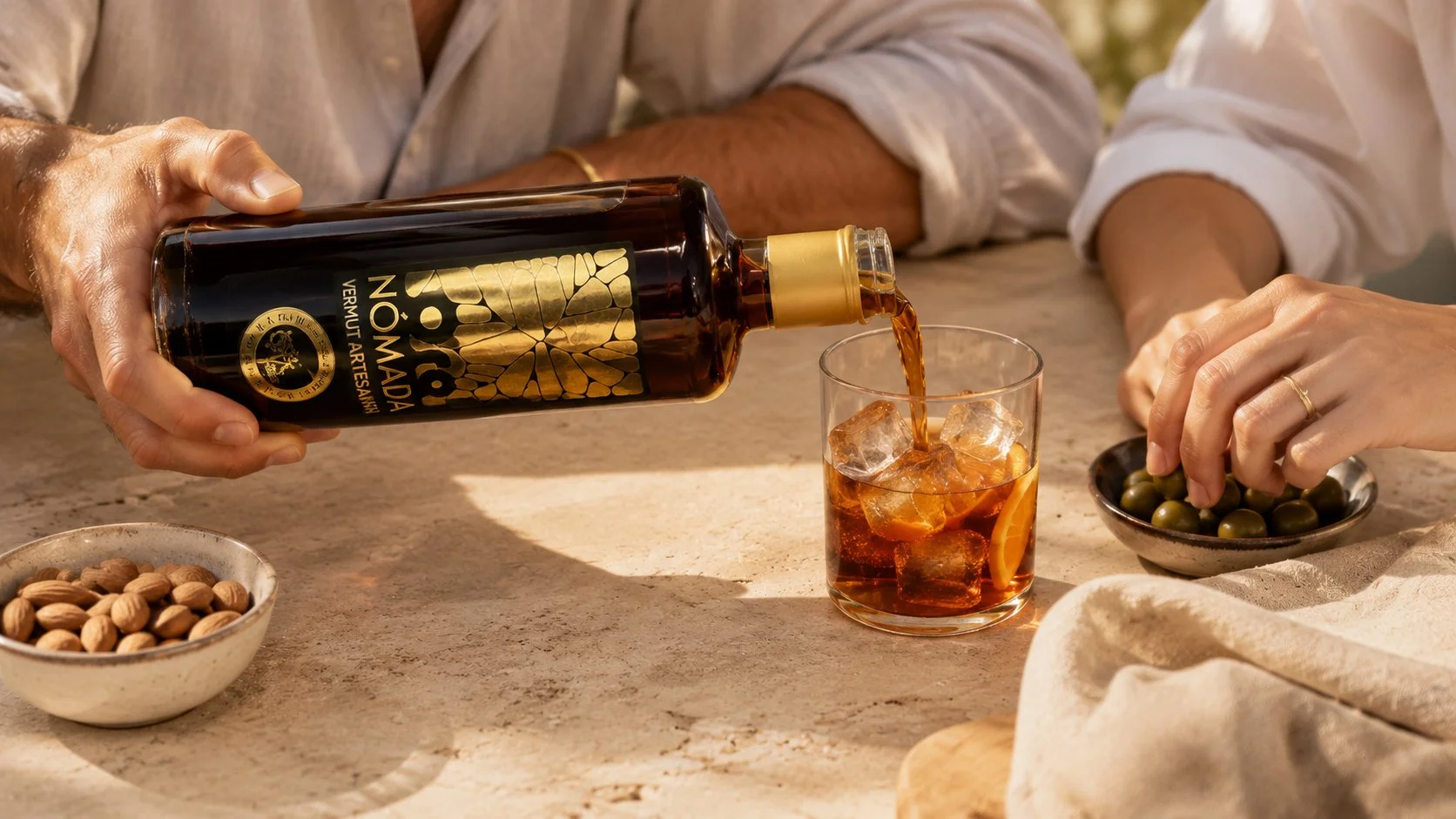 Persona sirviendo una bebida oscura Oro Nómada Vermut Artesano con hielo en un vaso, acompañado de almendras y aceitunas en platitos, en una mesa de piedra con un fondo neutro.