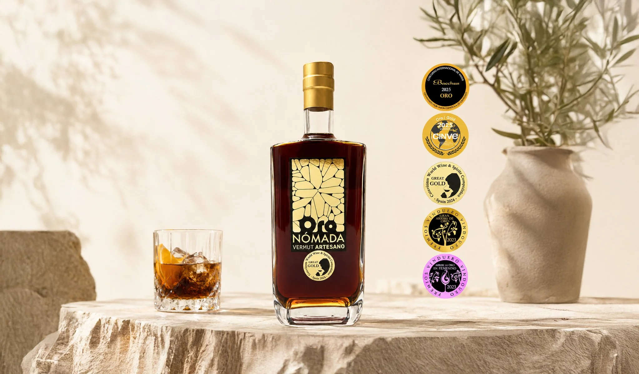 Botella de vermut artesanal Oro Nómada, con varios premios en etiquetas a su lado, acompañado de un vaso de mezclado con hielo y rodajas de naranja, decorado con una planta en un jarrón en un entorno luminoso.