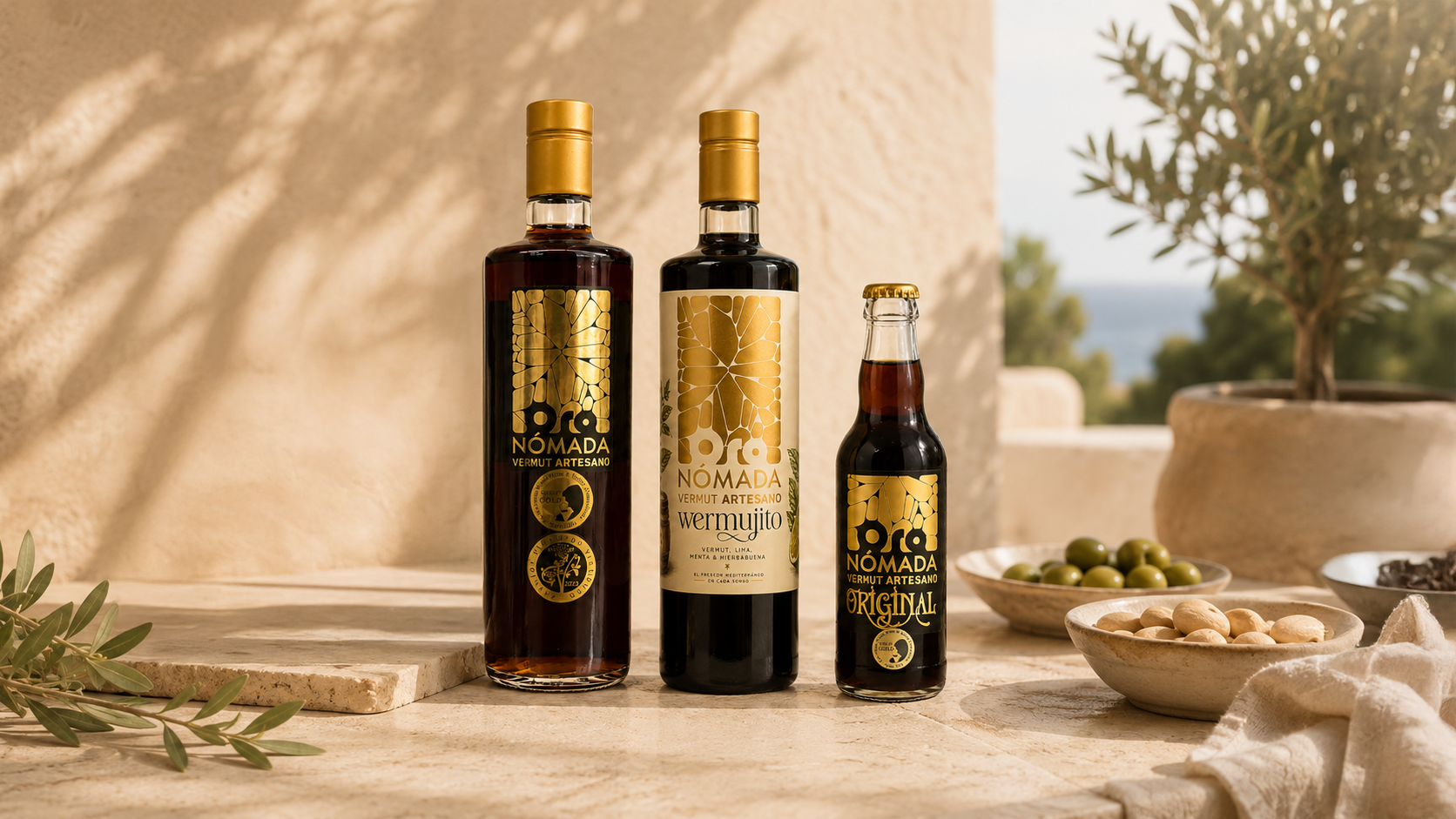 Tres botellas de vermut artesano oro nómada con etiquetas doradas y negras en una mesa de piedra, acompañadas de platos con aceitunas y frutos secos, en un entorno al aire libre con plantas y vista al mar.