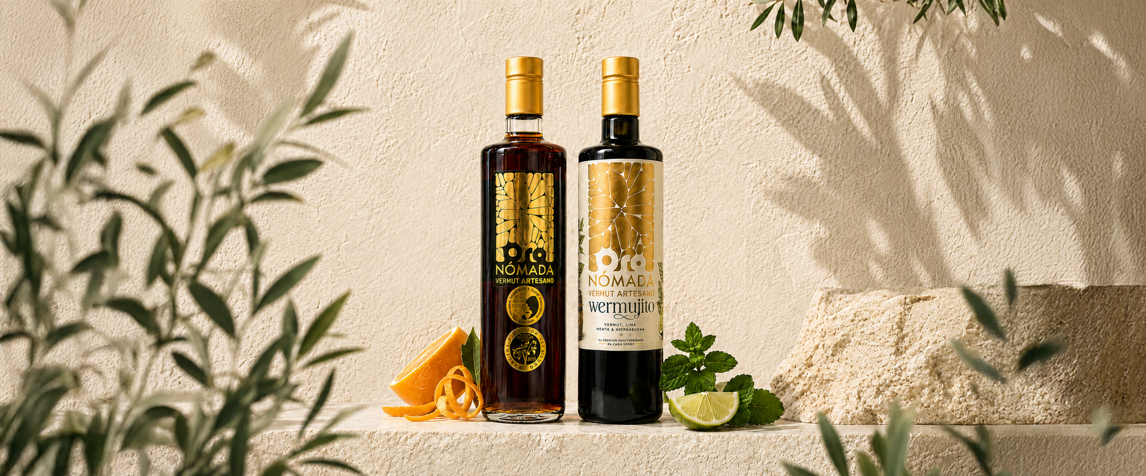 Dos botellas de vermut artesanal de la marca Oro Nómada en un fondo de pared beige, rodeadas de limón, menta, cáscara de naranja y hierbas, con un bloque de queso y hojas de plantas frondosas en primer plano.