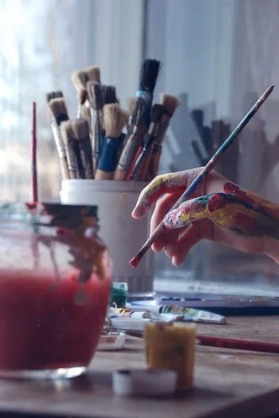 A person's hand covered in paint hovers above their workstation, which has painting supplies strewn about.