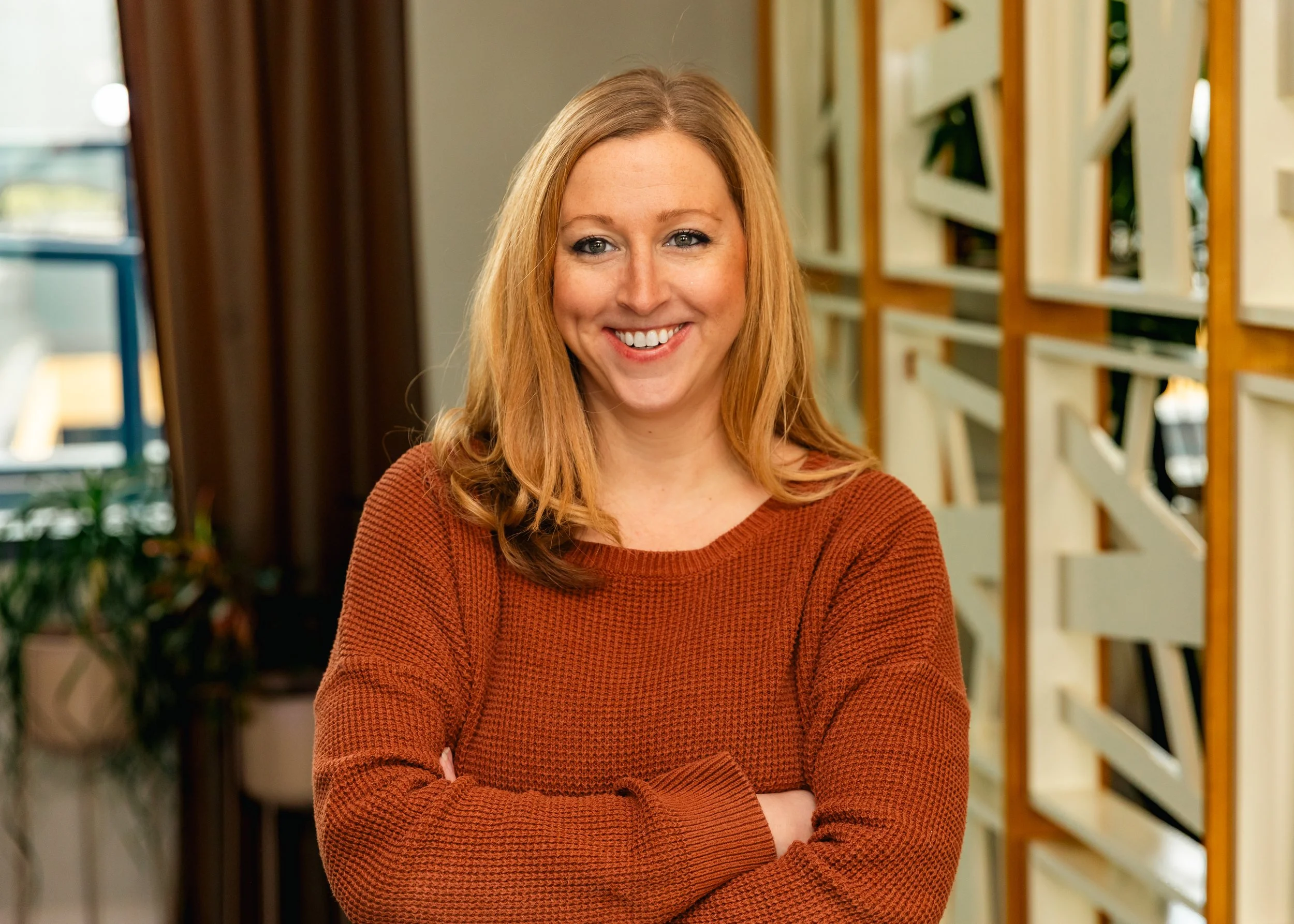 Woman with long blonde hair smiling in a cozy kitchen setting with brick wall background and decorative shelves.