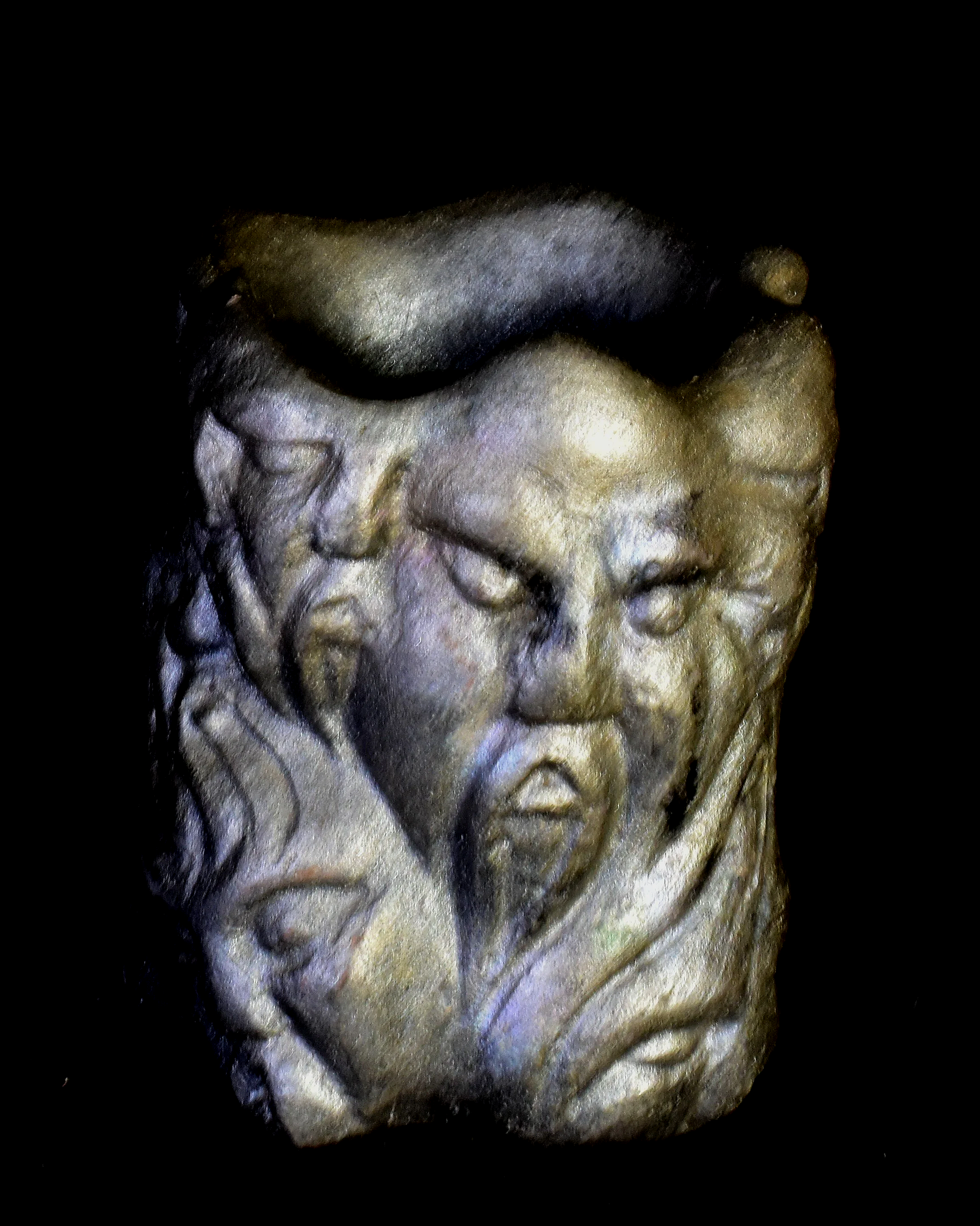 Close-up of a detailed stone or ceramic sculpture of a face, showing the forehead, eyes, nose, and upper cheeks, with hair carved in an ornate style, set against a black background.