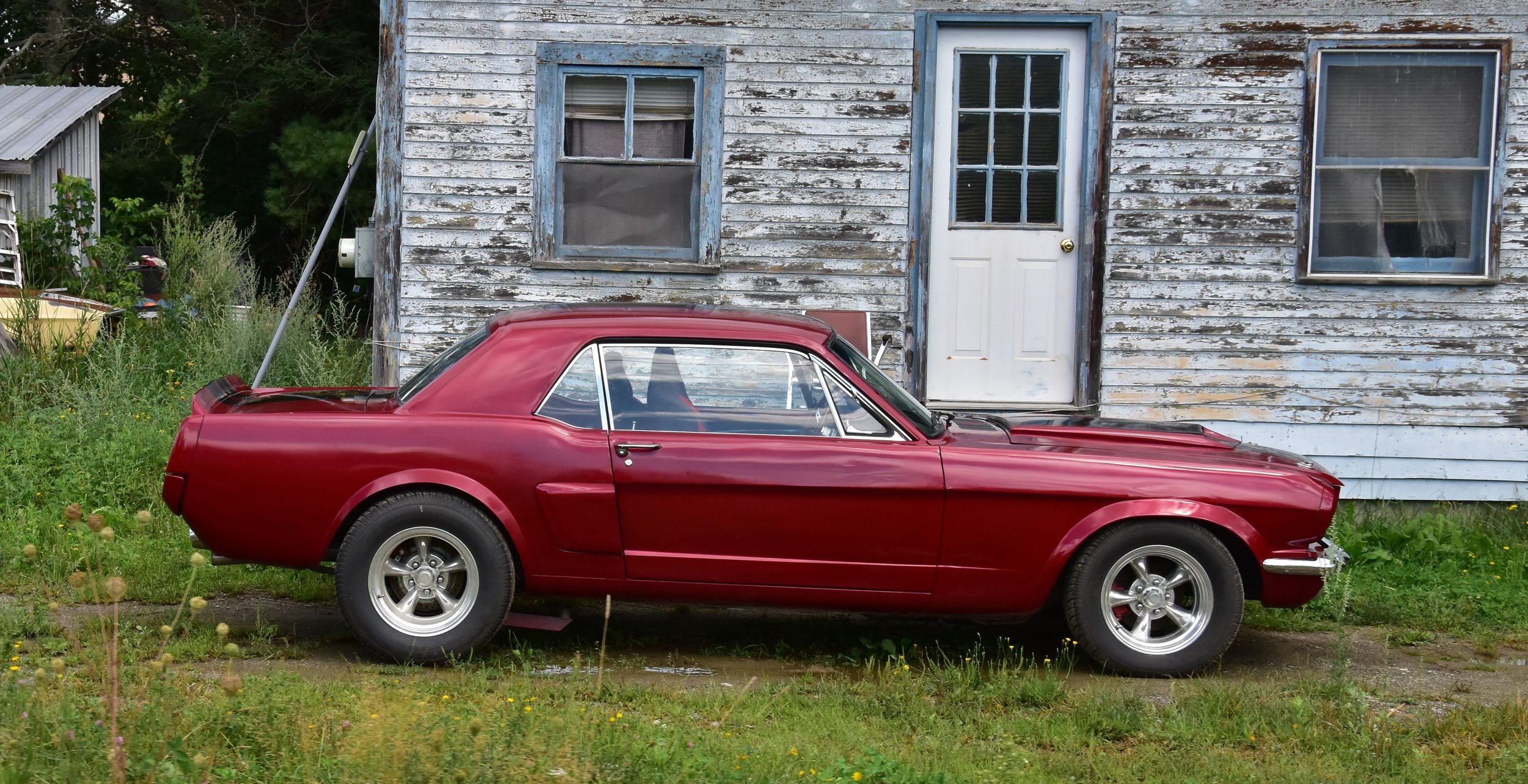 A red vintage sports car parked in front of a weathered, white wooden house with peeling paint, two windows, and a white door, surrounded by overgrown grass and plants.