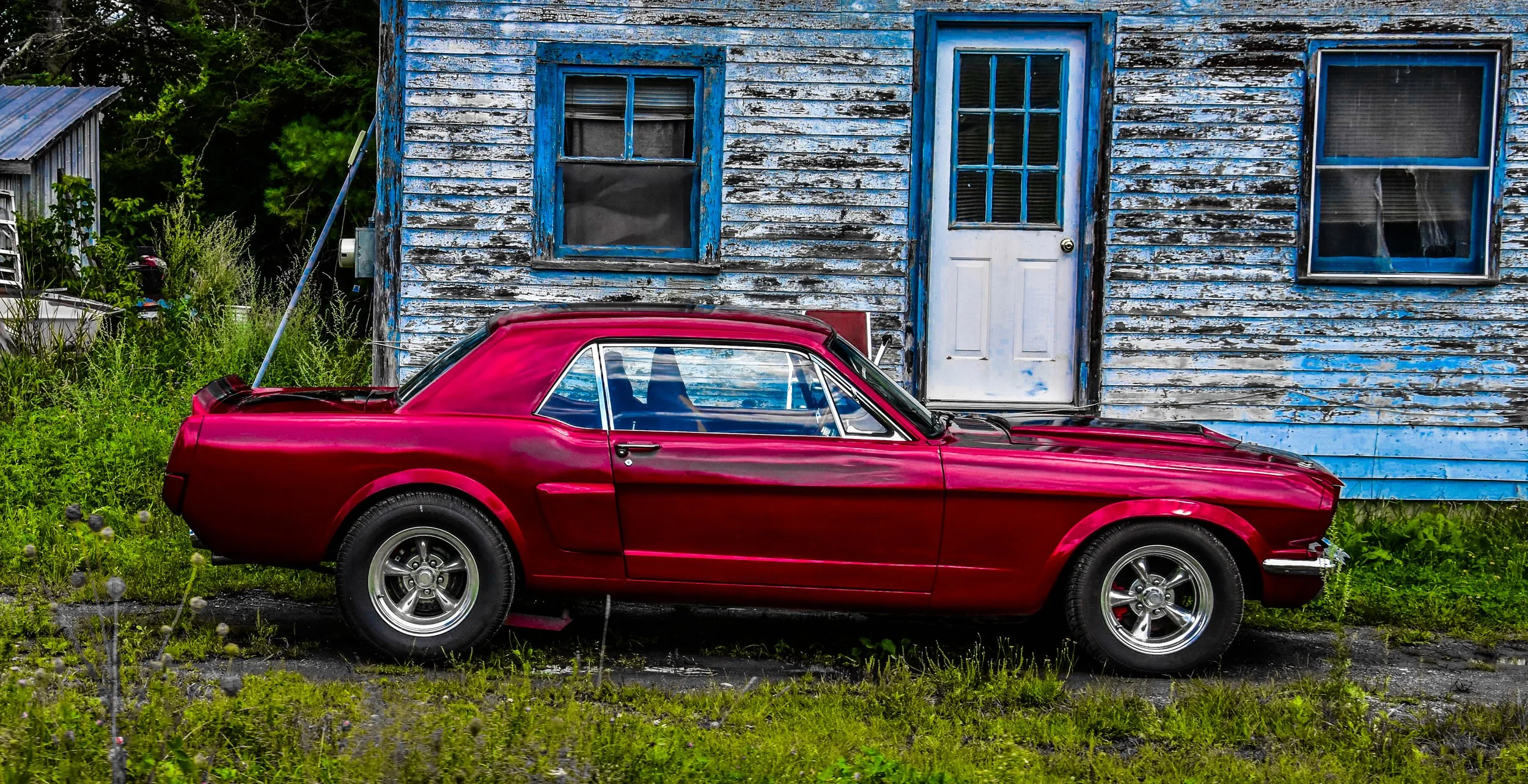 A red vintage sports car parked in front of a weathered blue wooden house with peeling paint and three windows.