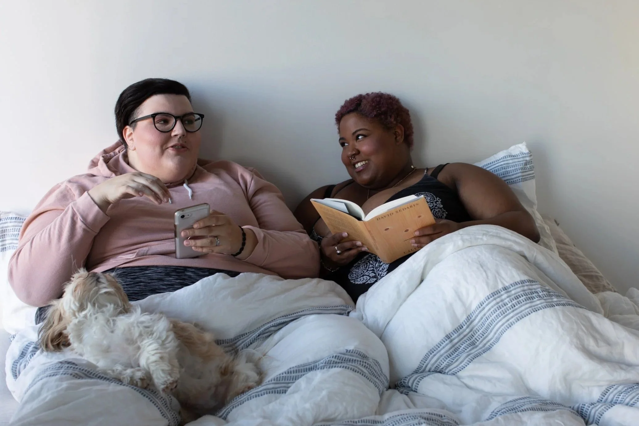 Two people sitting in bed, one holding a phone and the other reading a book, with a small dog lying on the bed.