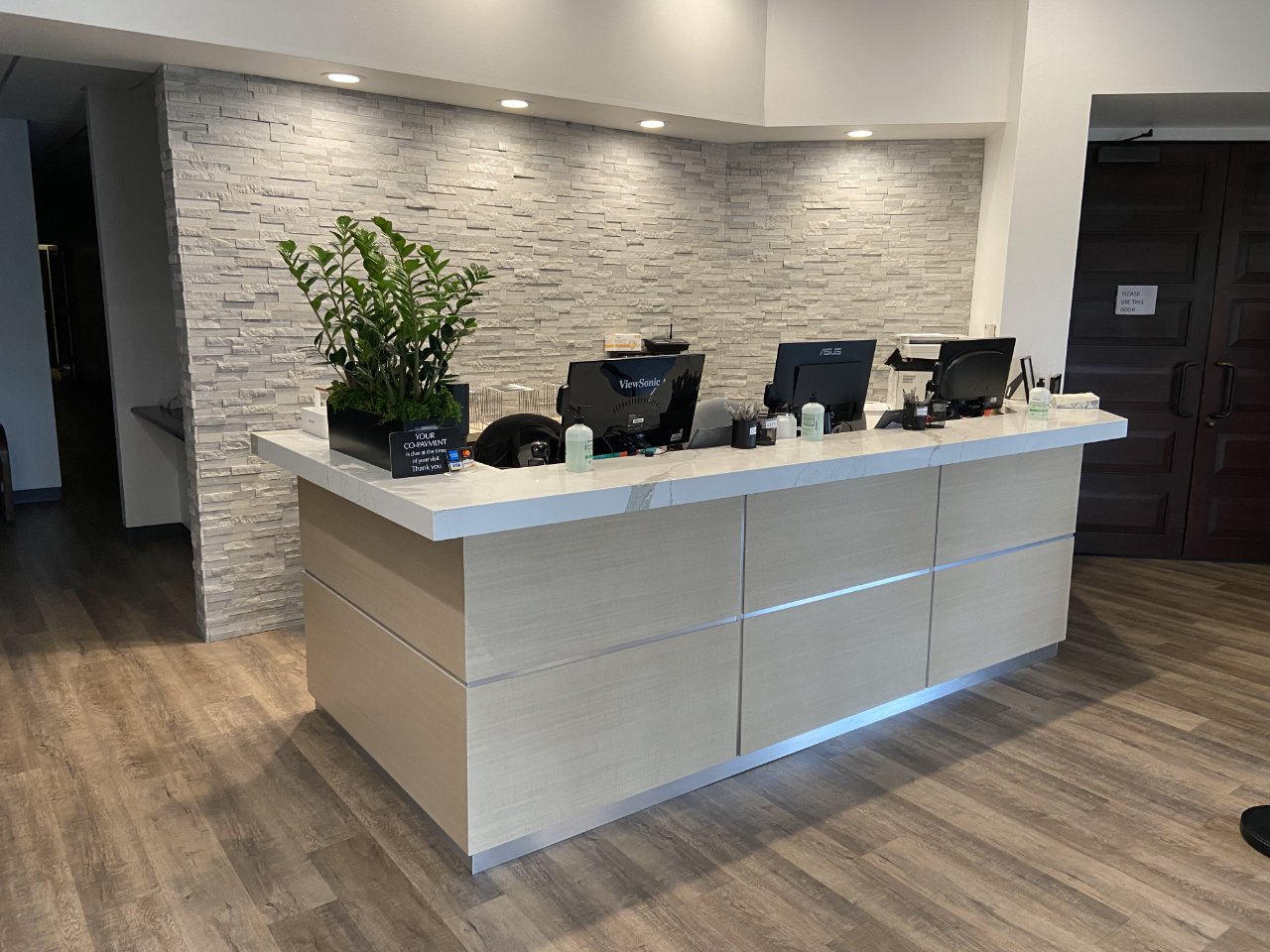 Custom made reception desk for a Dr's Office with stone wall backdrop, plants, and computer monitors.