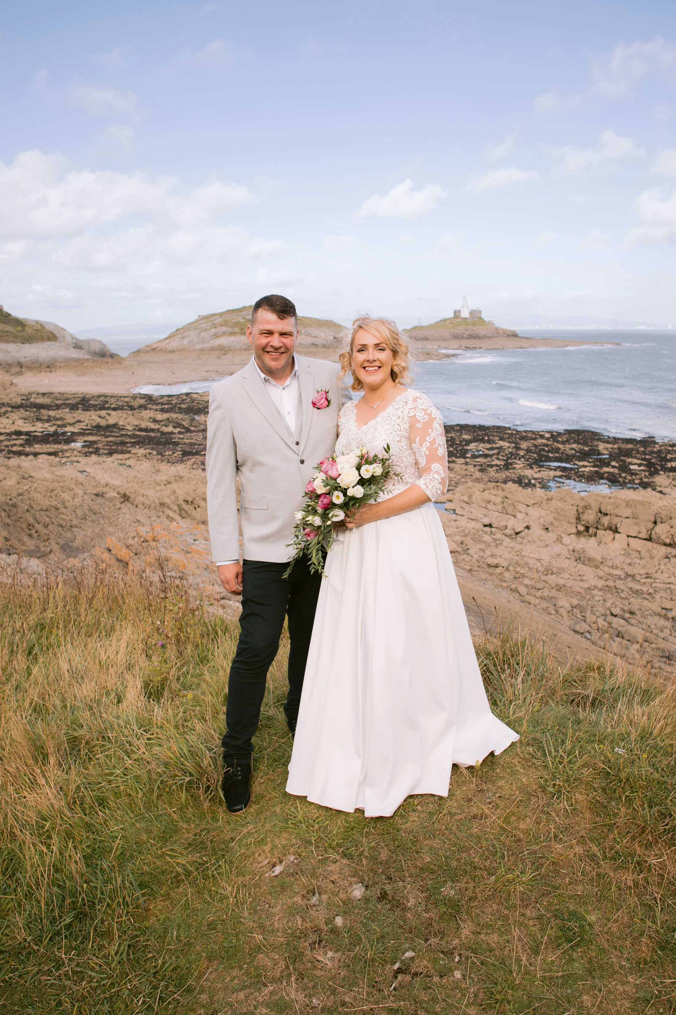 A newlywed couple standing on a grassy hill near the coast, with the groom in a light grey suit and the bride in a white wedding dress holding a bouquet, against a scenic ocean background with rocky cliffs and a lighthouse in the distance.