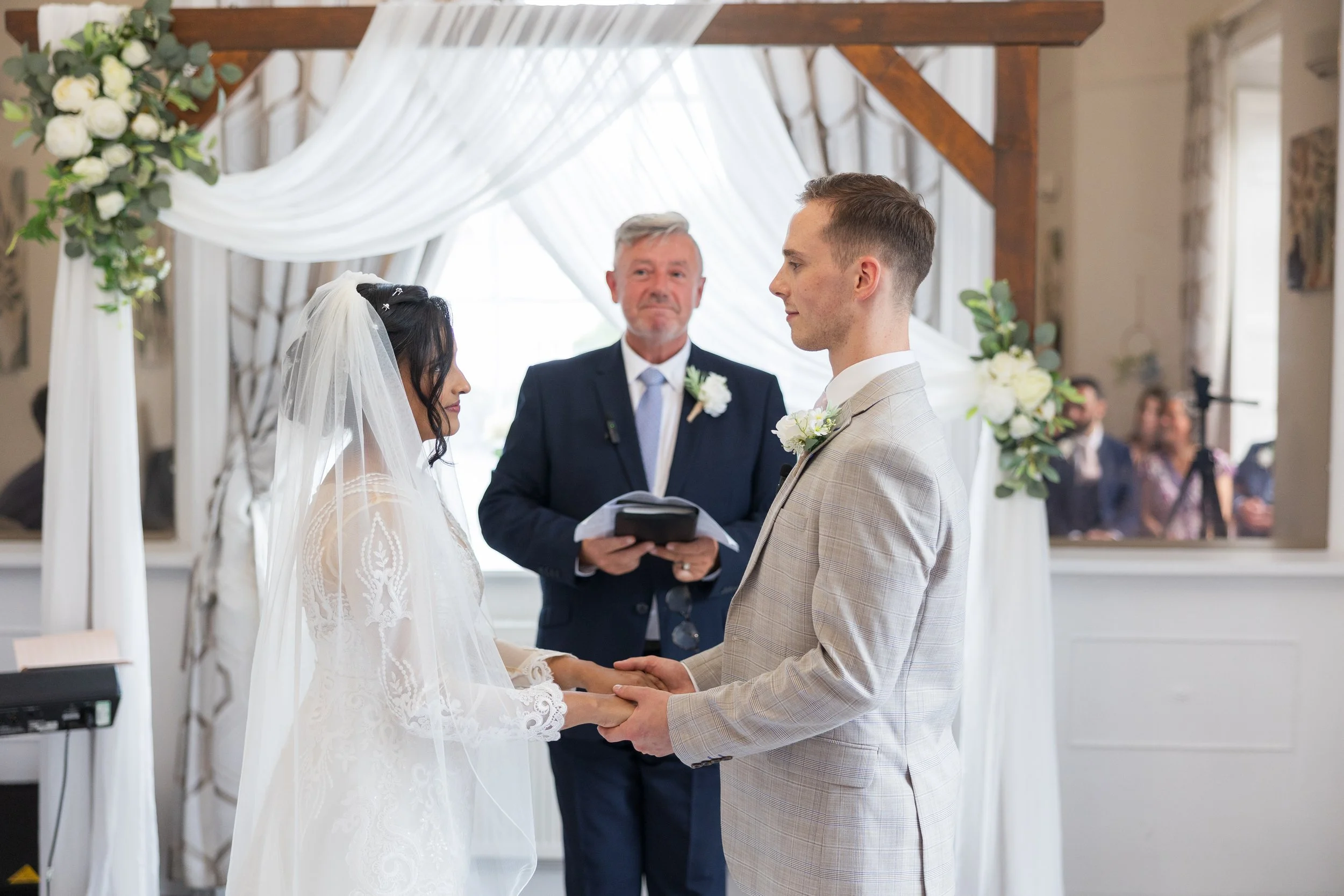 A bride and groom facing each other during their wedding ceremony, holding hands, with an officiant standing behind them, inside a decorated wedding venue with white drapes and floral arrangements. Norton House, Swansea.