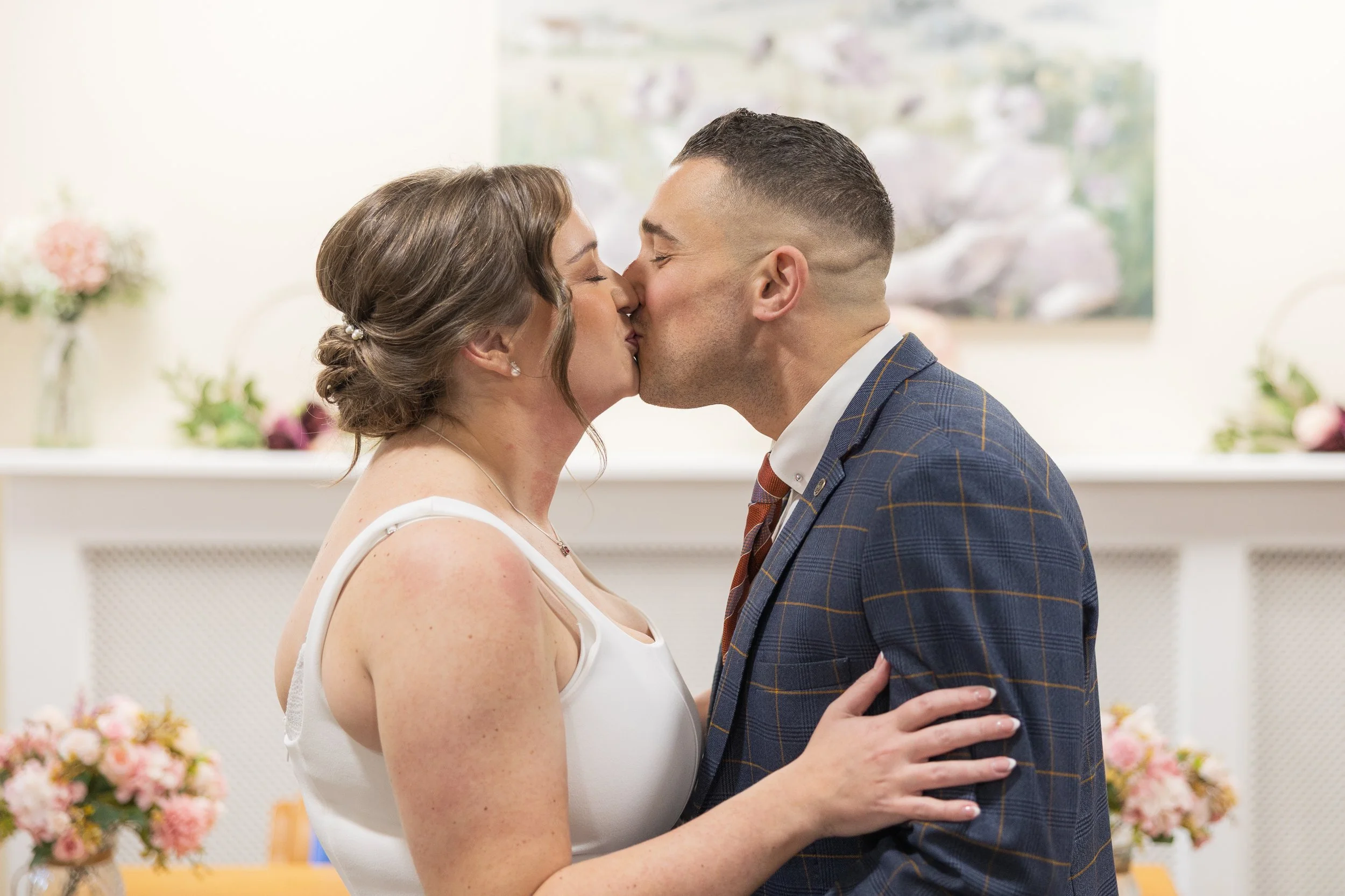 A couple is kissing at their wedding celebration, with flowers and wedding decor in the background. Neath Registry Office.