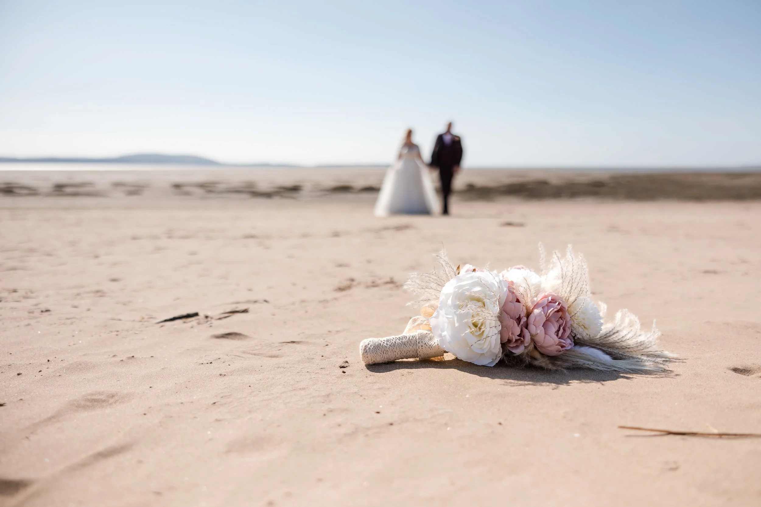 A wedding bouquet with pink and white flowers lying on sandy beach with a blurred couple in wedding attire in the background.
