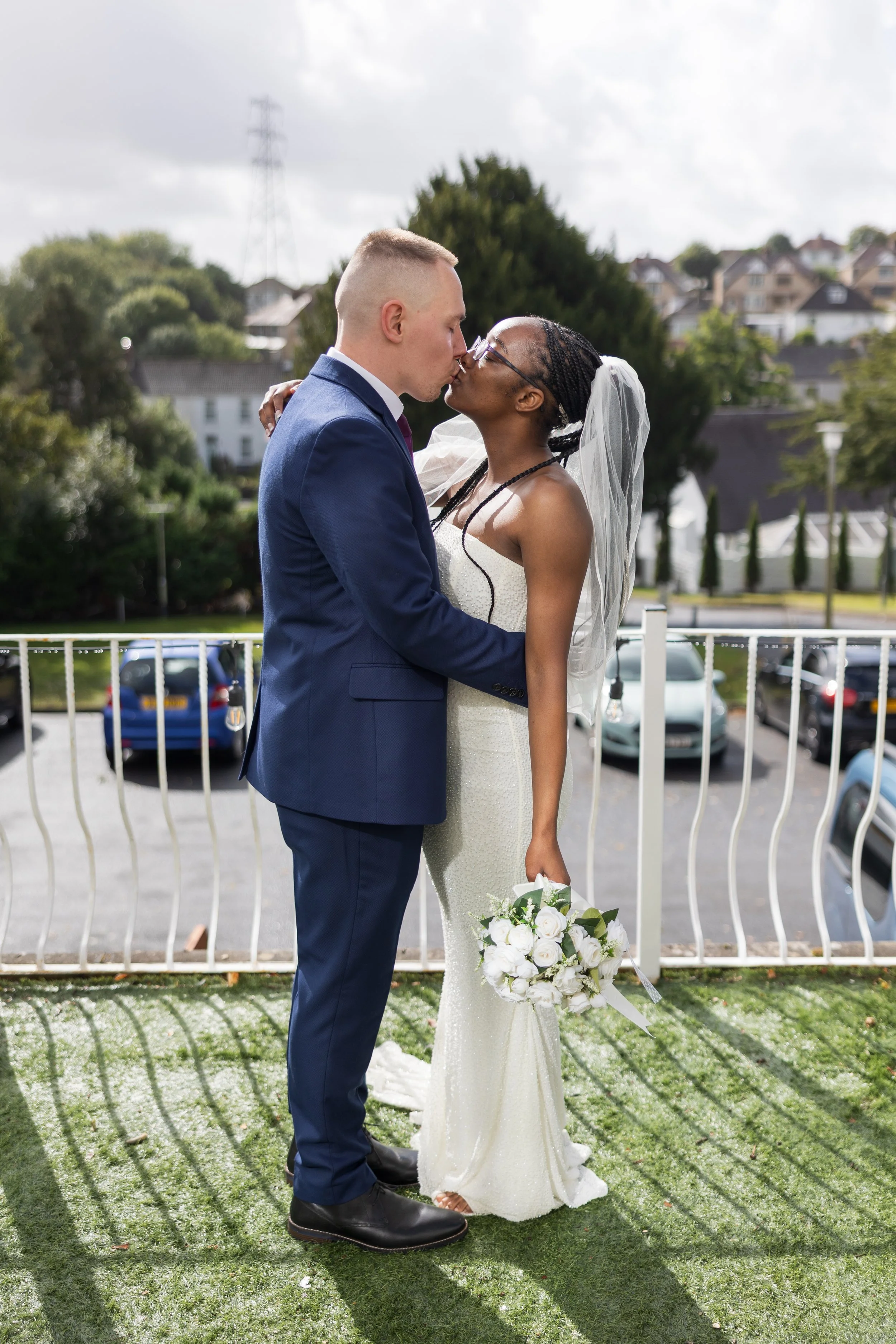 A newlywed couple shares a kiss on their wedding day, standing outdoors on a grassy balcony with a parking lot and residential neighborhood in the background.