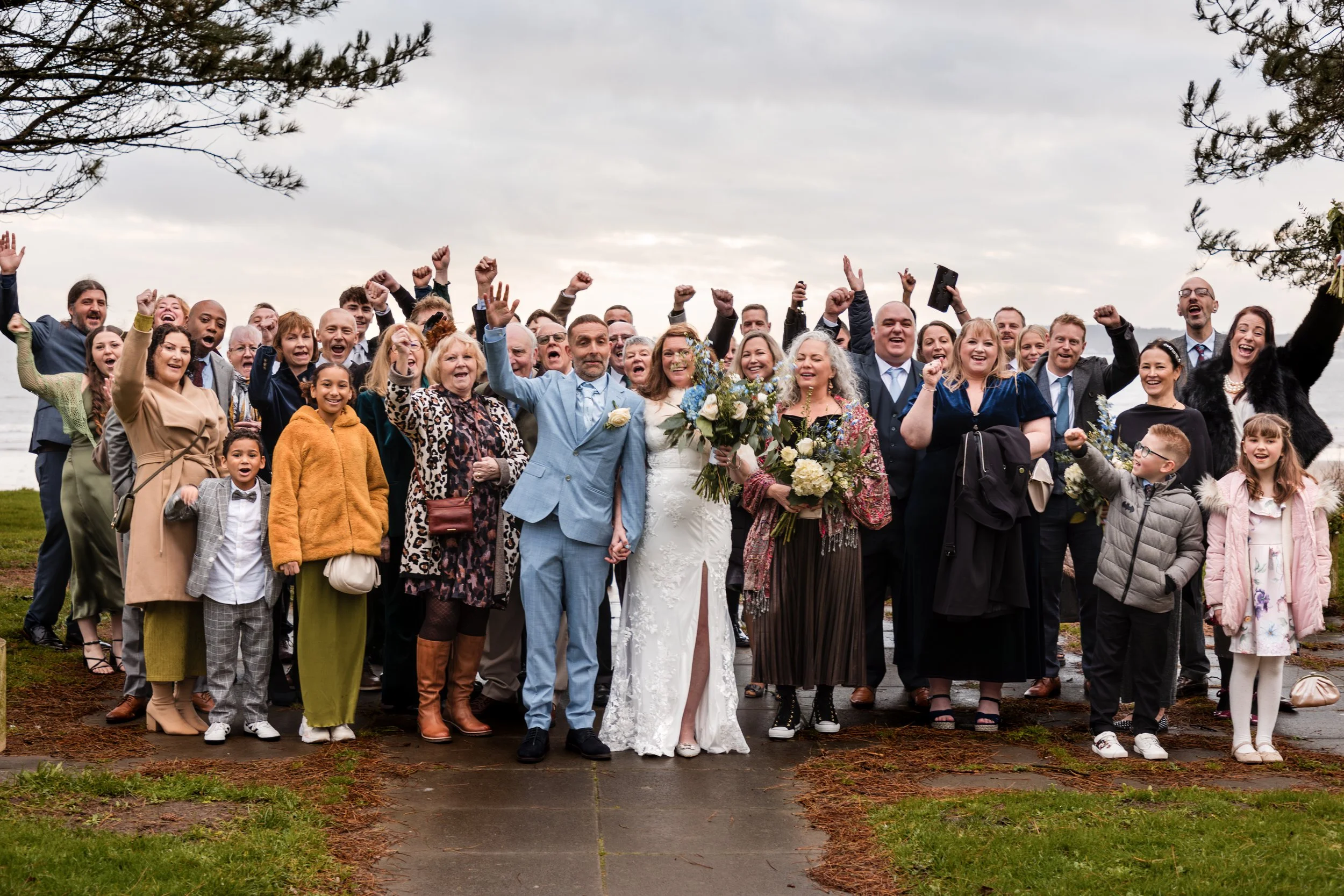 A group of people at a wedding outside, celebrating and smiling, with a cloudy sky and lake in the background. Swansea Registry Office.