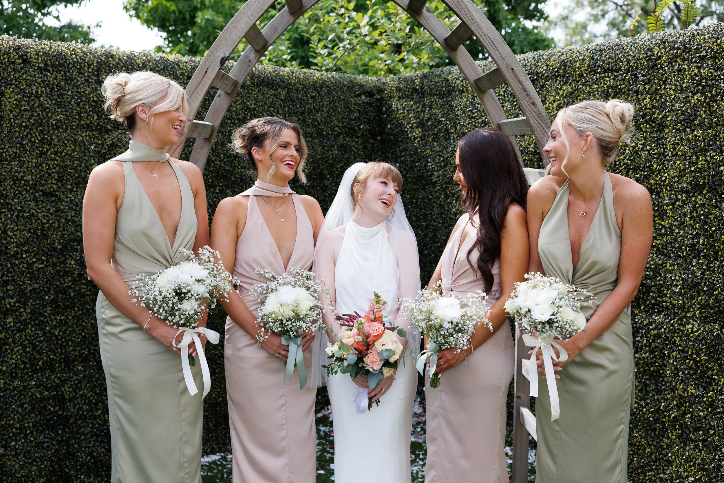 A bride and five bridesmaids standing under a garden arch during a wedding, all holding bouquets of flowers and smiling. Bridgend Registry Office.