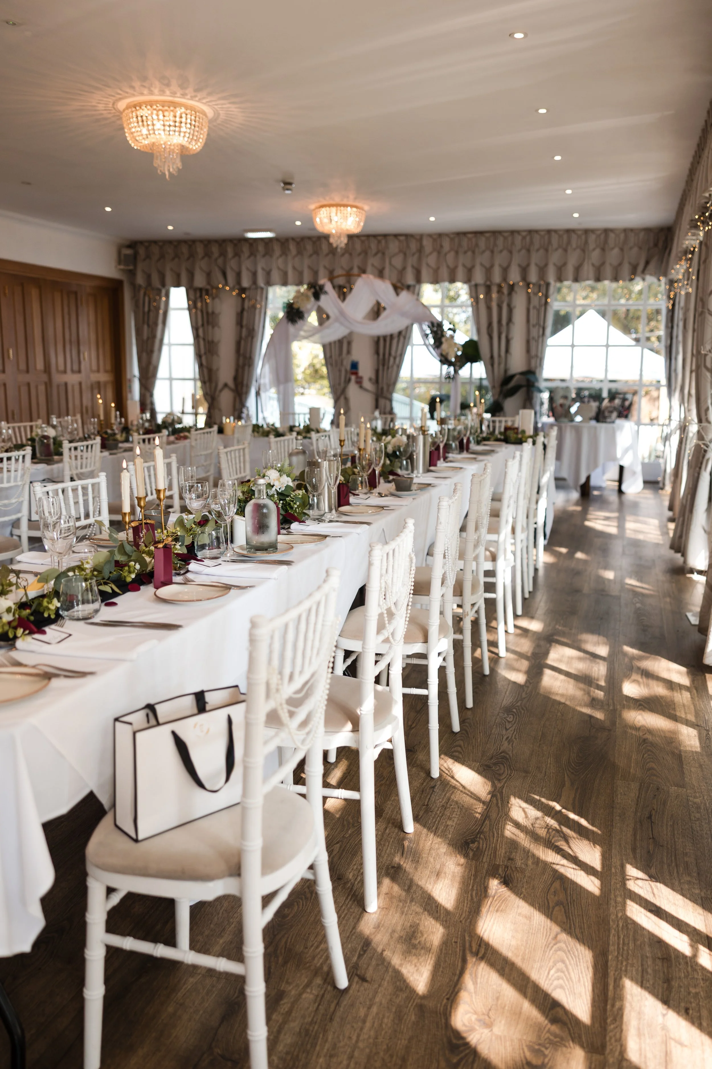 Elegant banquet hall set up for a celebration, with long white table, white chairs, floral centerpieces, candles, and natural light coming through large windows. Norton House, Mumbles.