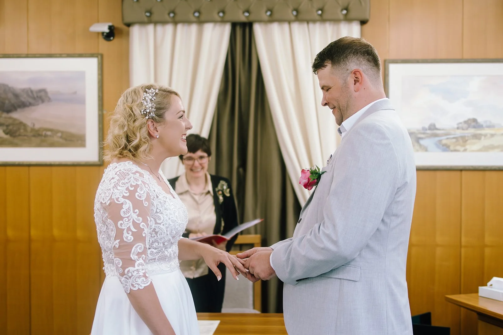 A bride and groom exchanging wedding vows indoors, holding hands and smiling at each other. The bride is wearing a white lace dress with floral details in her blonde hair. Swansea Registry Office.