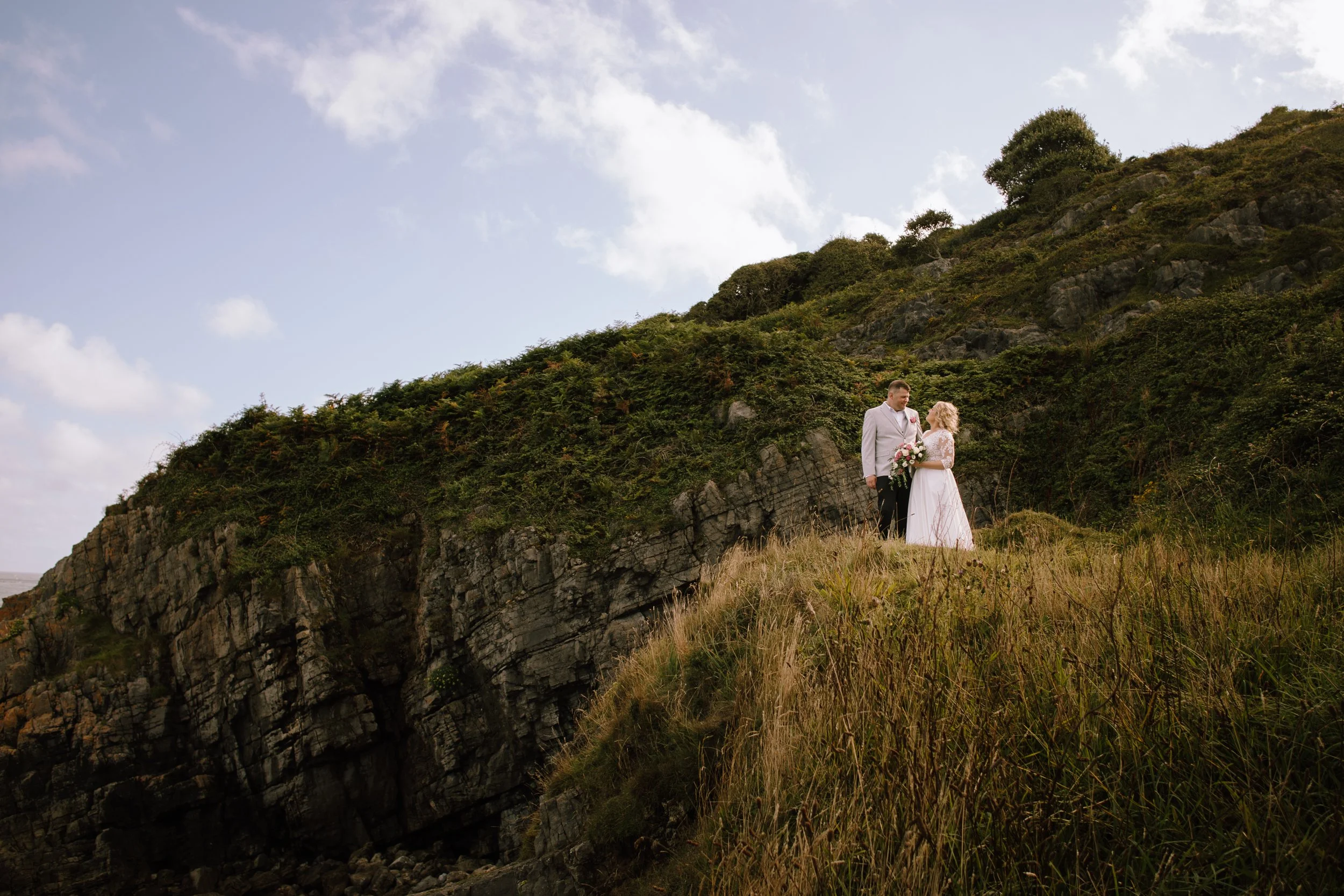 A bride and groom standing together on a grassy hill with rocky cliffs and a partly cloudy sky in the background.