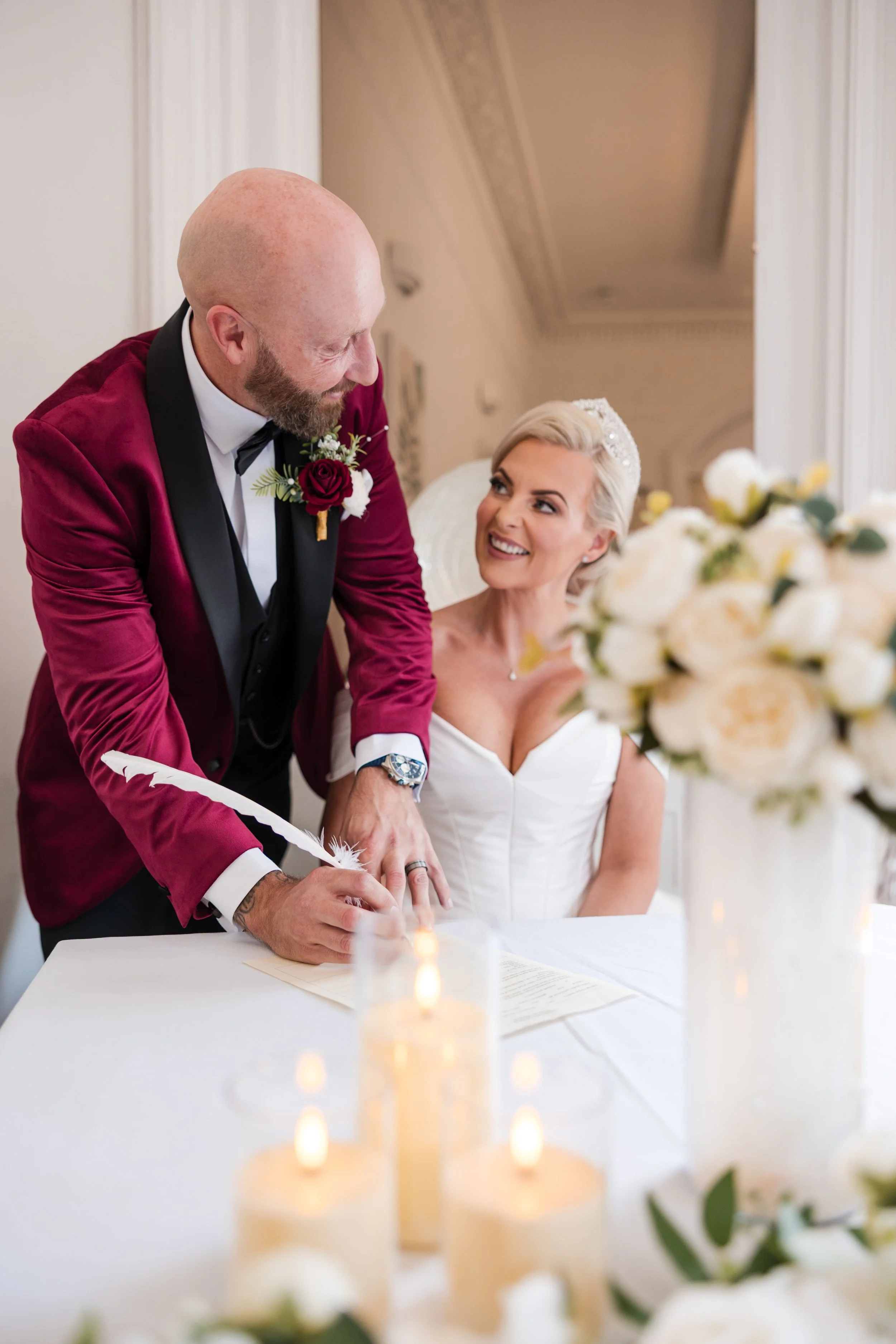 A groom in a red velvet tuxedo signing a marriage certificate while the bride, in a white wedding dress, looks at him smiling. The bride has platinum blonde hair and the groom is bald with a beard. Norton House, Swansea.