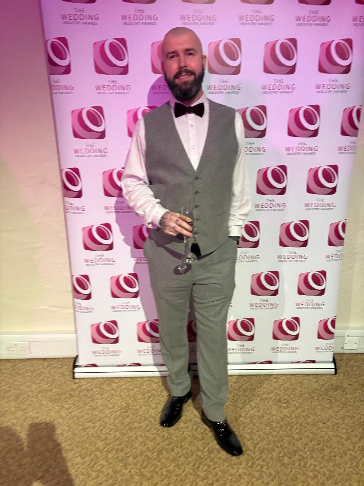 A man with a beard and tattoos, dressed in a white shirt, gray vest, plaid gray pants, black shoes, and a black bow tie, holding a champagne glass, standing in front of a pink and white step-and-repeat banner with the text 'The Wedding' and a pink logo.
