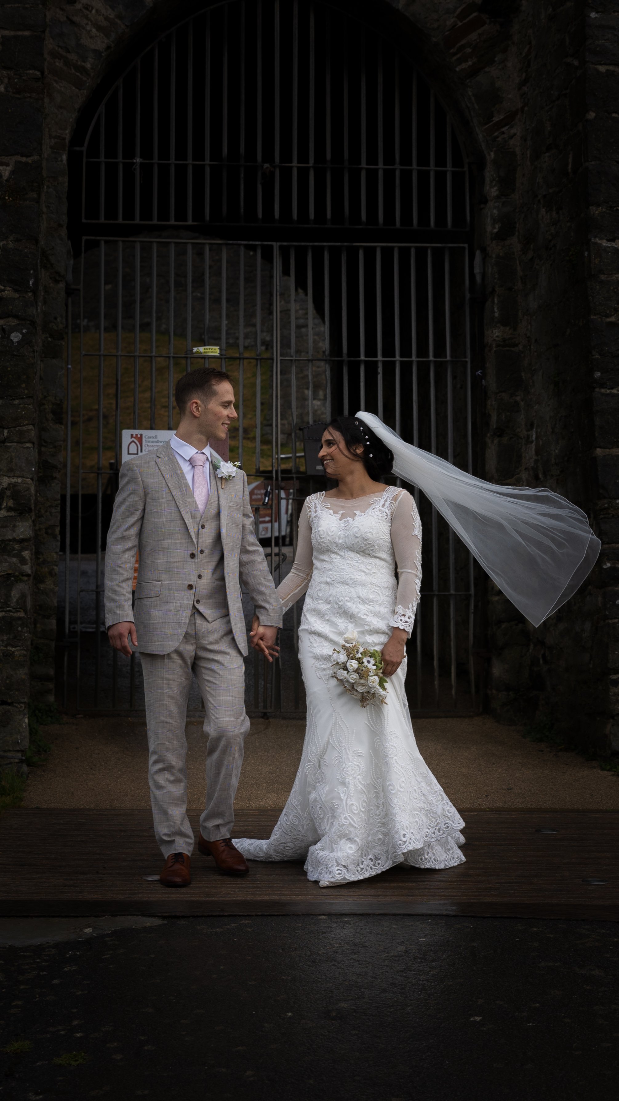 A bride and groom holding hands, standing in front of a large iron gate, smiling at each other during their wedding ceremony. Oystermouth Castle.
