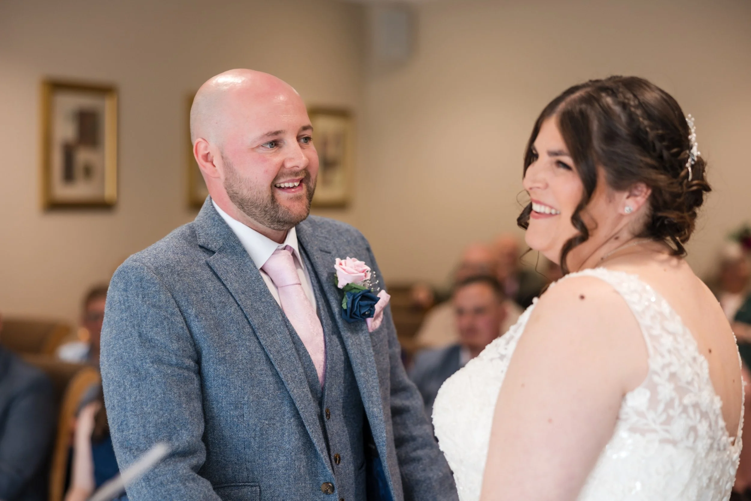 A groom and bride during their wedding ceremony. The groom is smiling and wearing a gray suit with a pink tie and a boutonnière, while the bride is smiling in a white lace wedding gown.