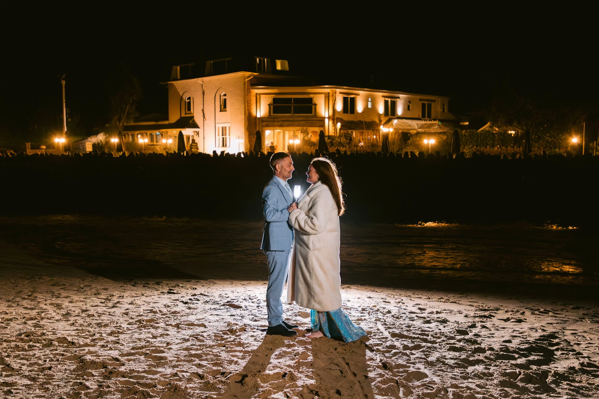 A couple dressed in formal attire standing on the beach at night, holding hands and gazing into each other's eyes, with a lit house in the background.