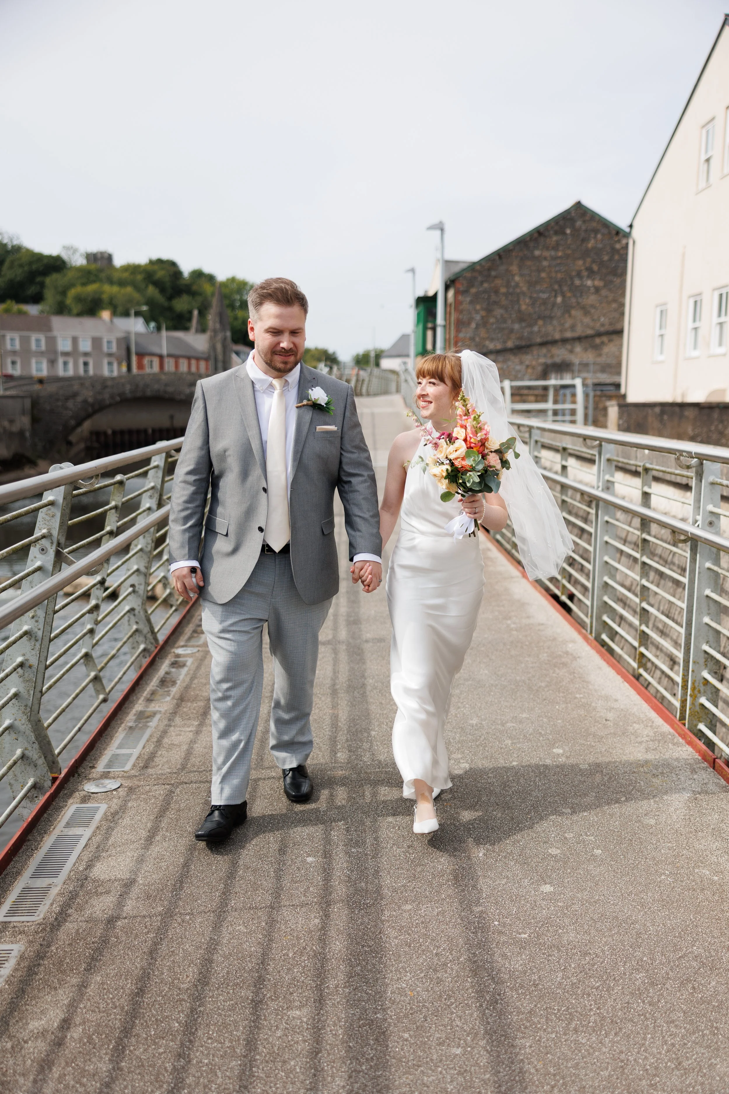A newly married couple walking hand in hand on a bridge, with the bride holding a bouquet of flowers.
