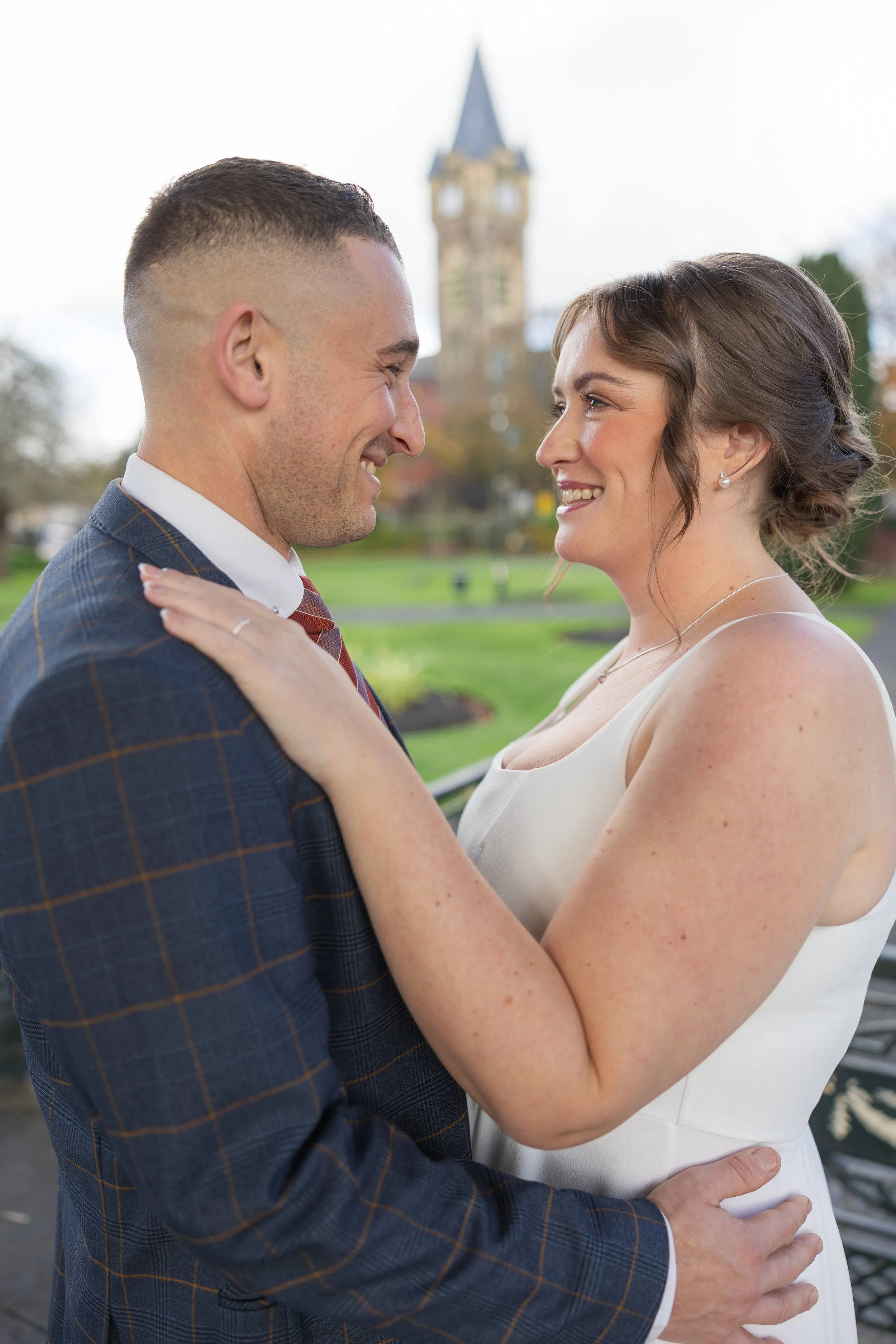 A couple embracing outdoors, smiling at each other, with a historic building with a clock tower in the background. Neath.