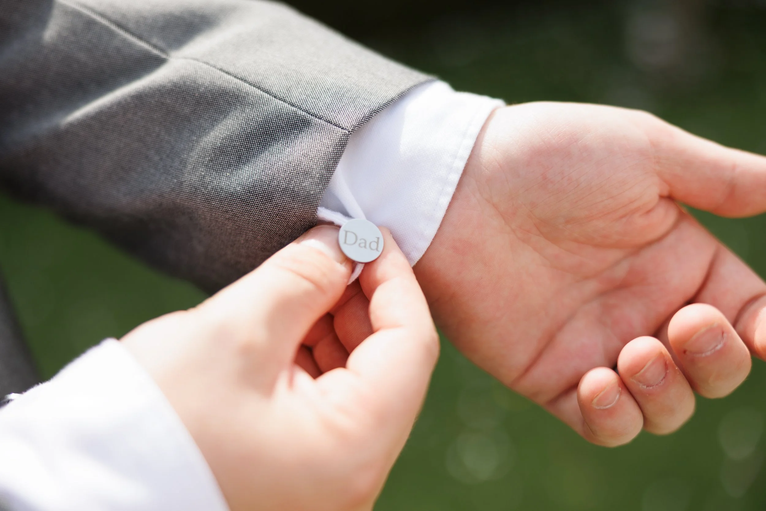 Close-up of a person in a suit with a white shirt, attaching a small, round, gray label with the word 'Dad' to their sleeve.