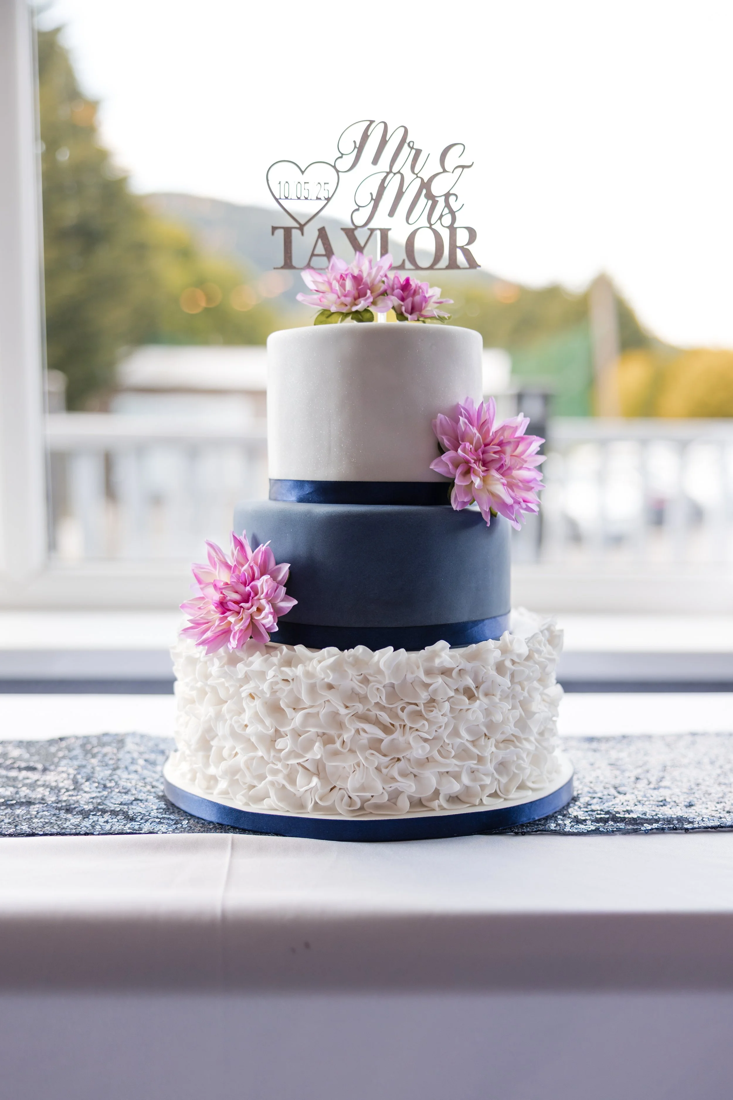 A three-tier wedding cake with white, navy blue, and decorated white ruffled layers, topped with pink flowers and a topper reading 'Mr & Mrs Taylor' with a heart and the date 10.05.25.