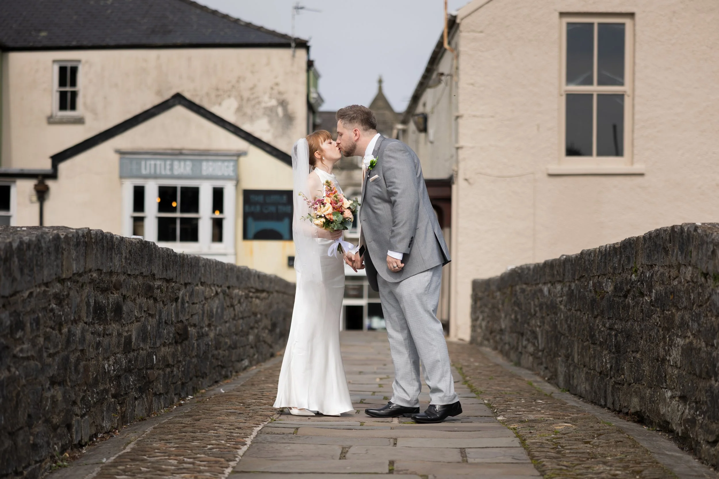A bride and groom share a kiss on a stone bridge in a small town, holding hands with the bride holding a bouquet of flowers.