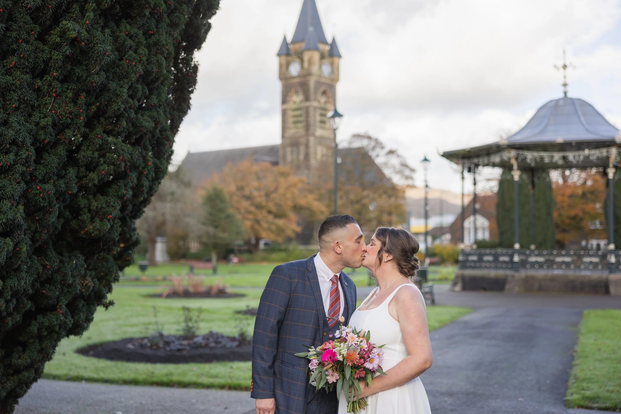 A couple in wedding attire sharing a kiss in a park with a church, gazebo, trees with fall foliage, and cloudy sky in the background. Victoria Gardens, Neath.