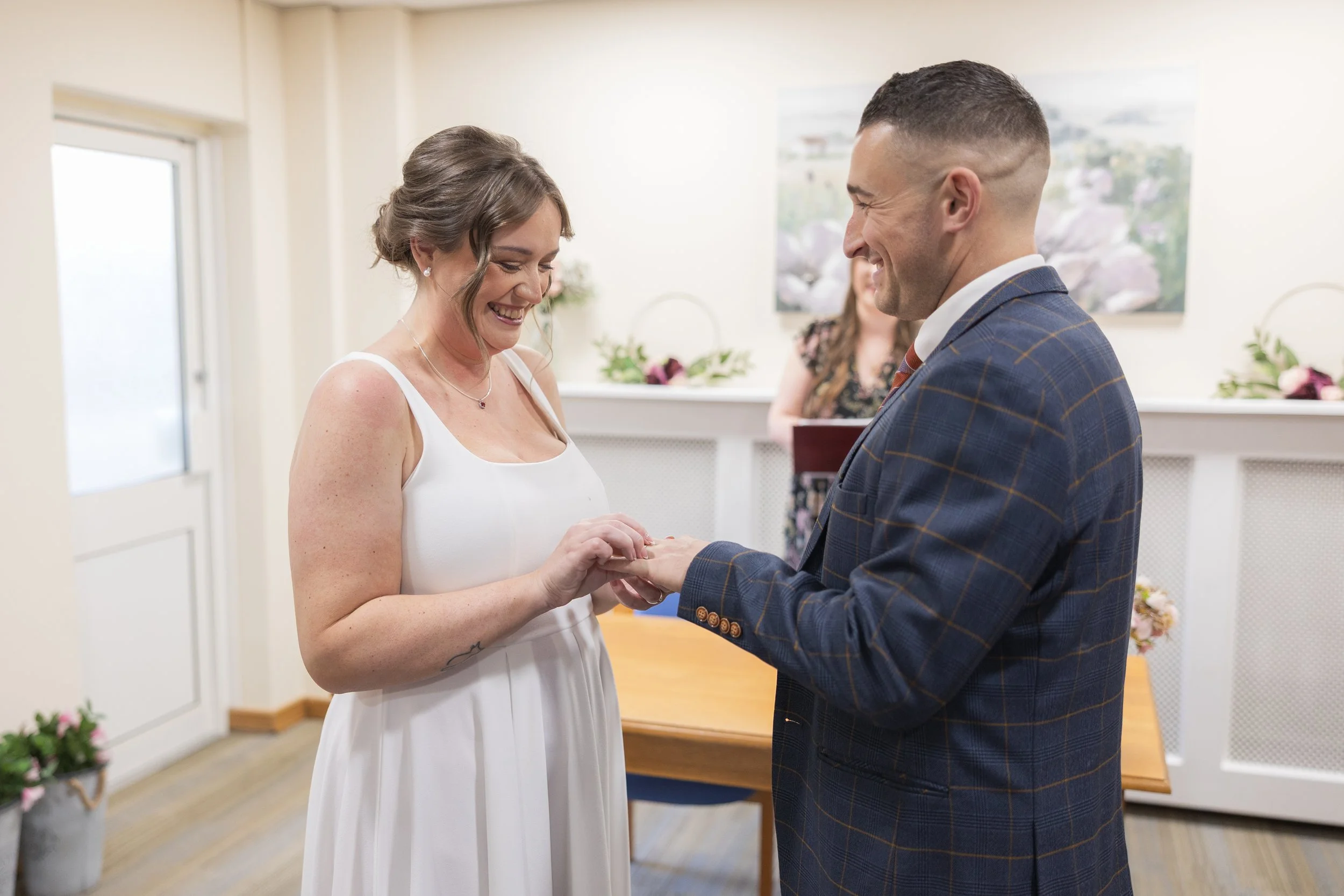 A couple, a woman in a white dress and a man in a checkered blazer, exchanging rings during a wedding ceremony in a bright room with floral decorations. Neath Registry Office.