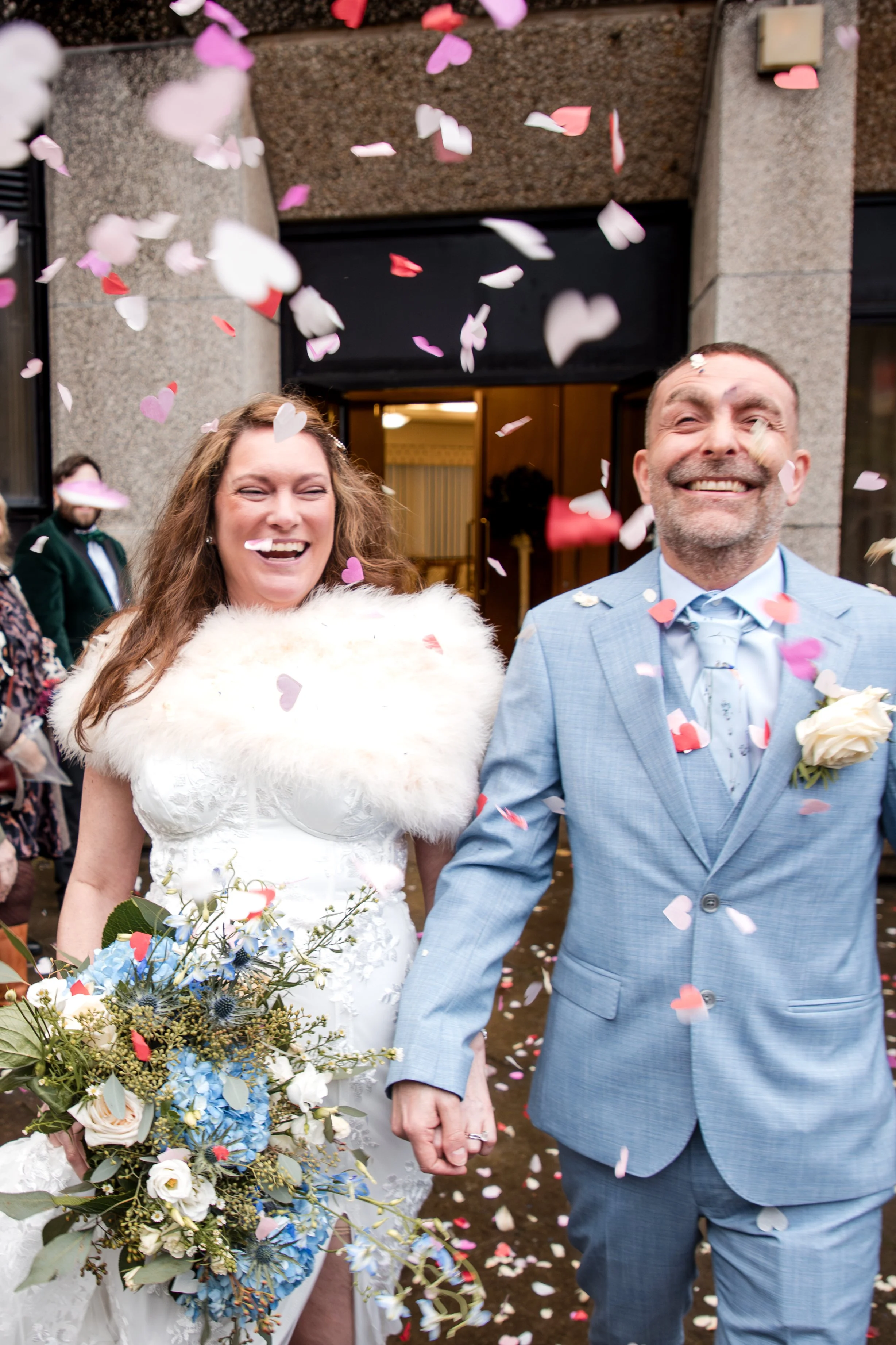 Happy bride and groom holding hands outside, surrounded by falling confetti at their wedding celebration. Swansea Registry Office.