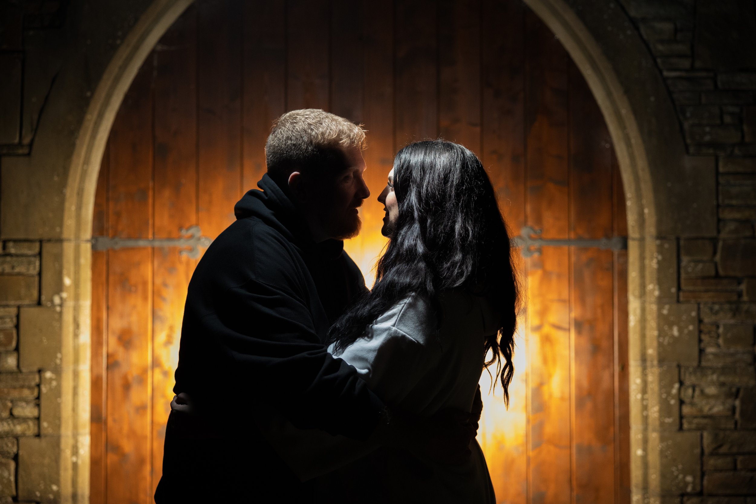 A couple with dark hair and light hair facing each other, close together indoors with a warm wooden door behind them, in a romantic moment at The Glyn Clydach, Neath.