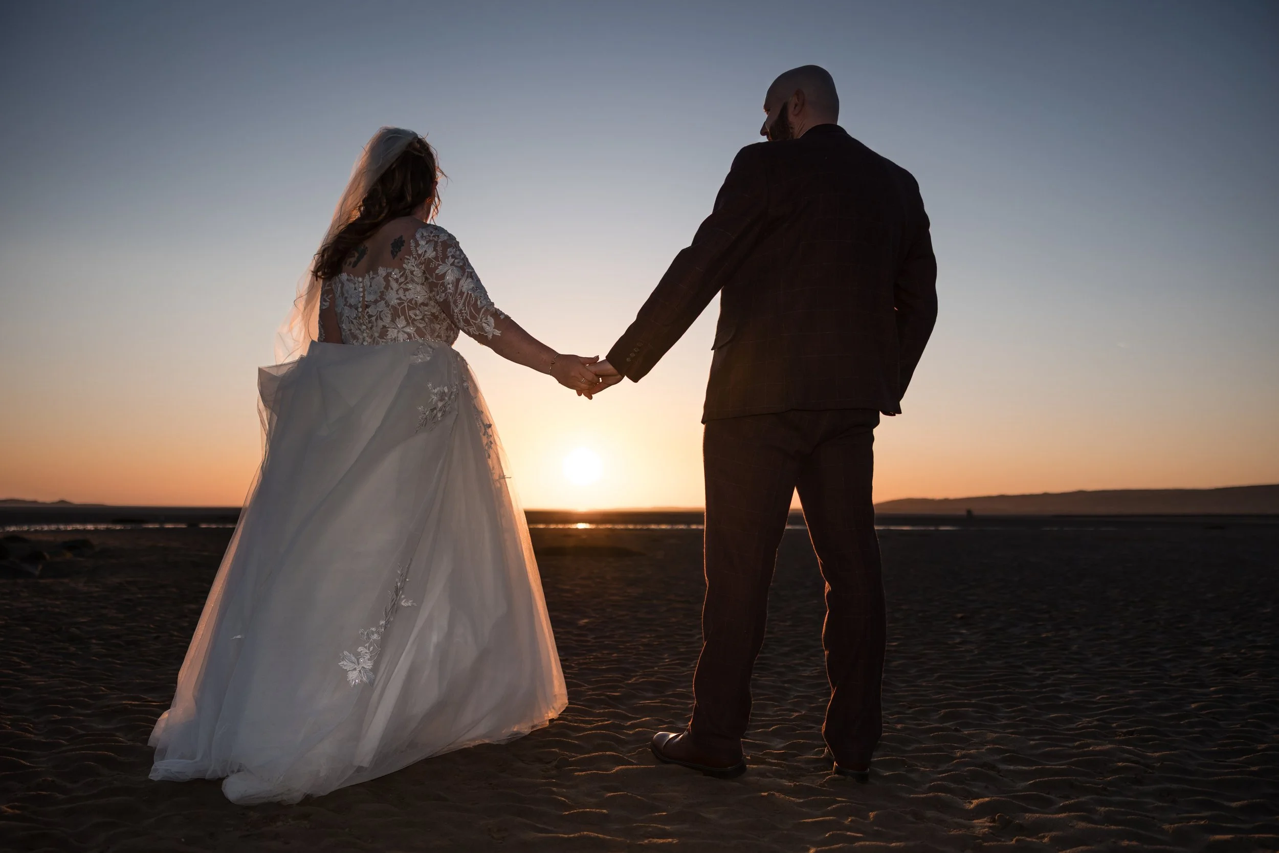 A bride and groom holding hands at sunset on a beach, with the bride in a white wedding gown and the groom in a dark suit.