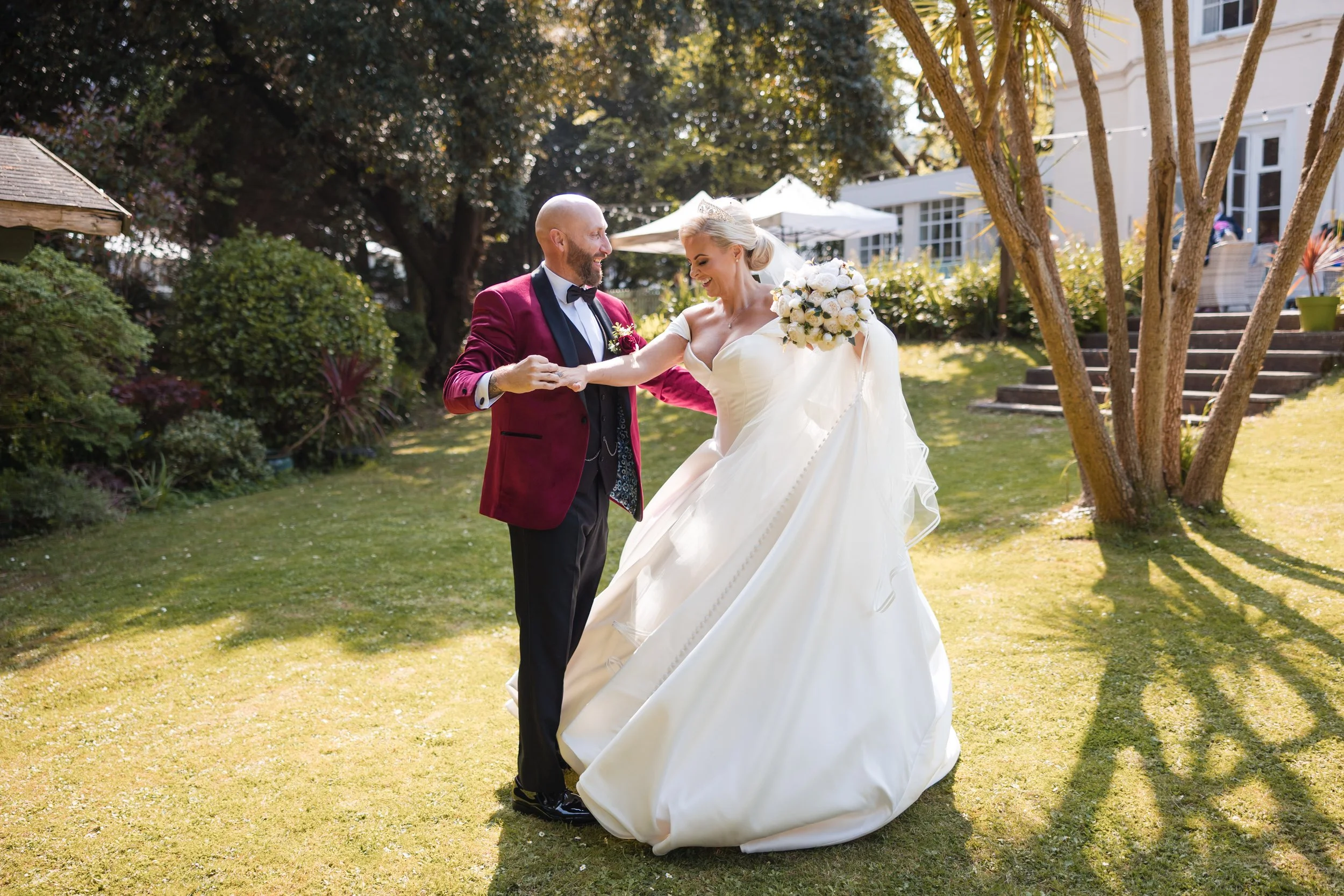A newlywed couple dancing outdoors on a sunny day, with the bride wearing a white wedding gown and holding a bouquet of white flowers, and the groom in a red jacket, black pants, and a bow tie, surrounded by greenery and trees. Norton House, Mumbles.