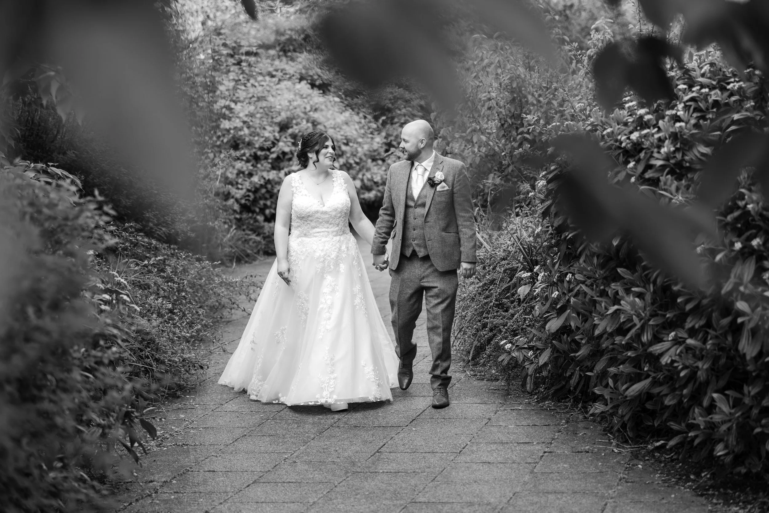 Black and white photo of a bride and groom walking hand in hand on a garden path, looking at each other, surrounded by lush foliage.