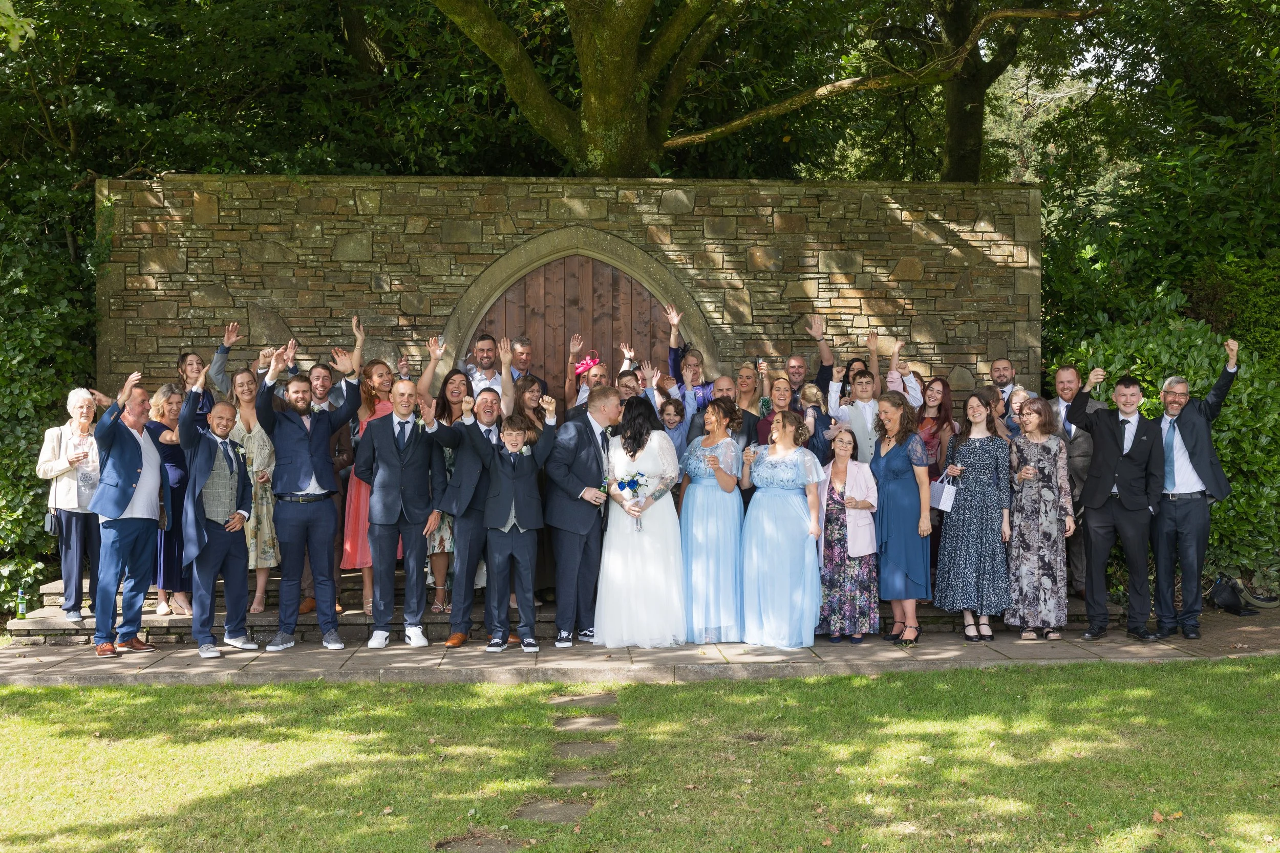 A large group of wedding guests celebrating outdoors in front of a stone wall with a wooden arched door, waving and smiling; the bride and groom are in the center, with the bride holding a bouquet. Glyn Clydach, Neath