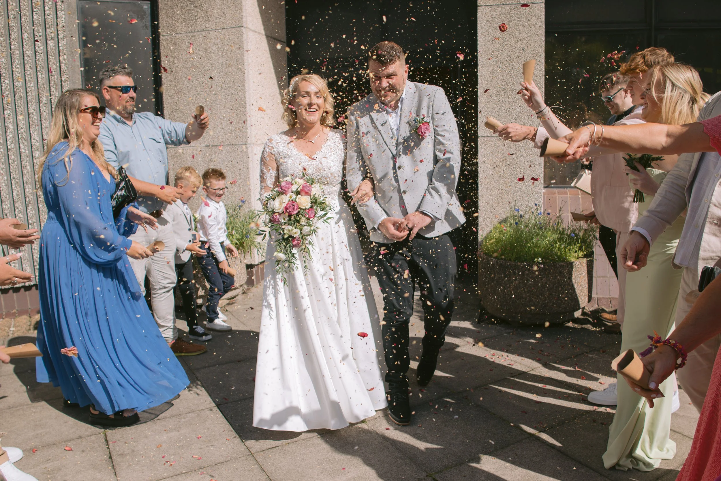 A newly married couple walking through a confetti celebration outside a building, surrounded by friends and family. Swansea Registry Office.