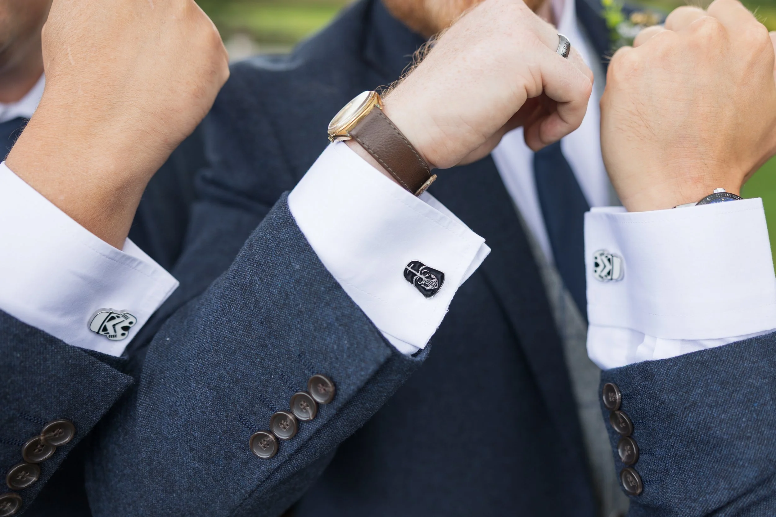 Two men in suits showing their cufflinks and flexing their arms, taken outdoors.