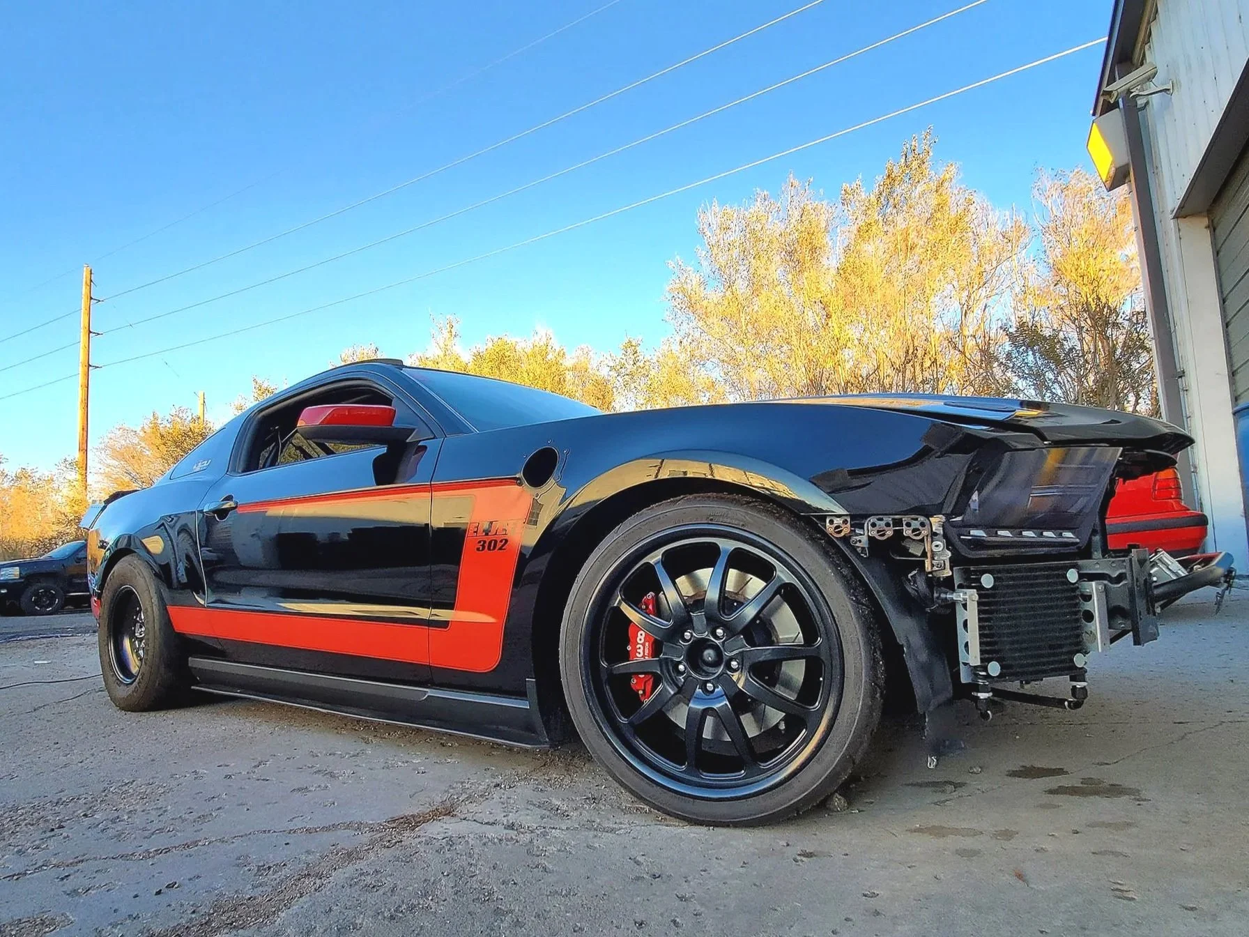 A black sports car with red accents and a partially disassembled front end parked outside near a warehouse and other vehicles, under a blue sky with trees in the background.