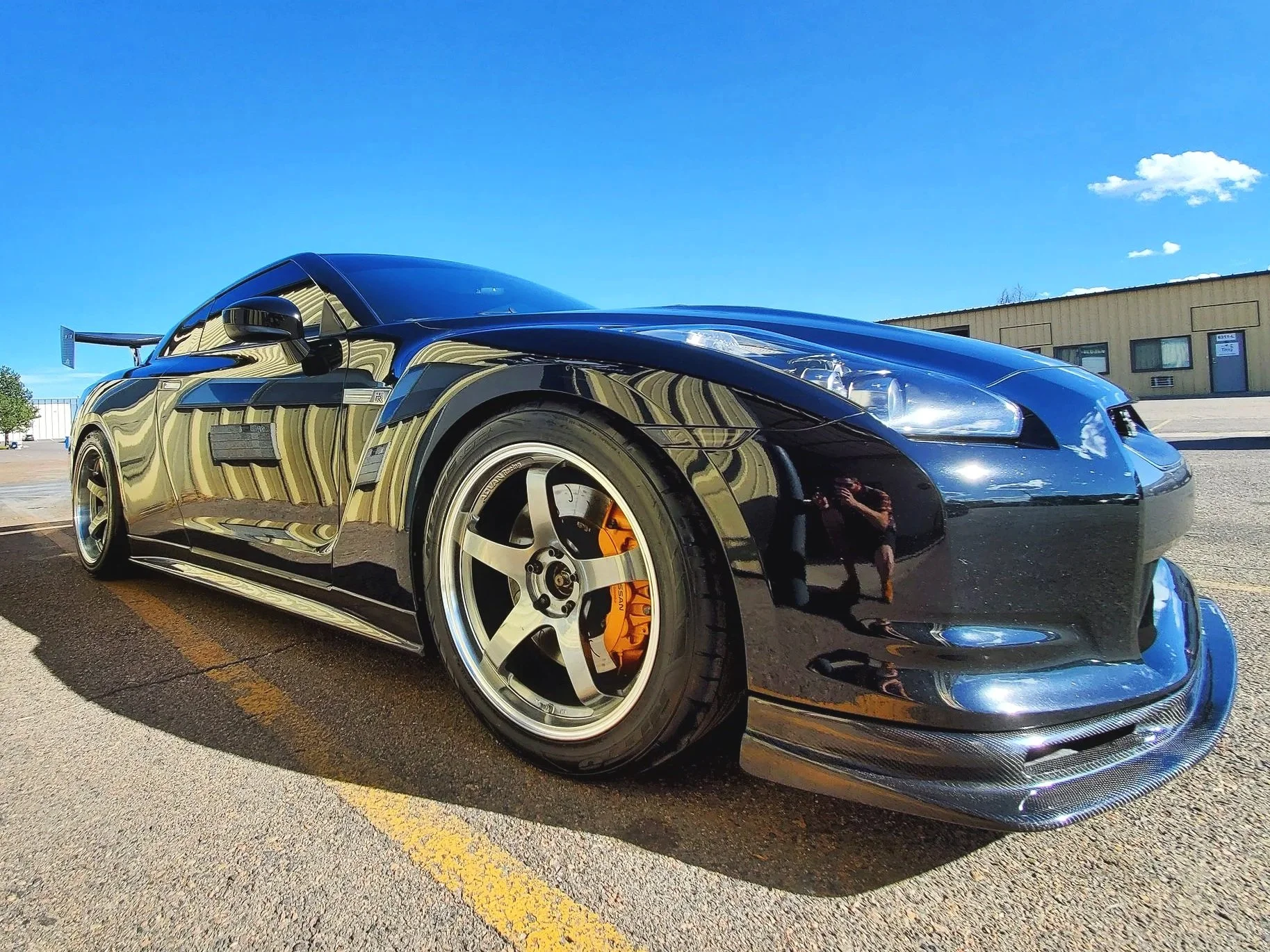 A black sports car GTR with aftermarket wheels, orange brake calipers, and a large rear wing is parked outdoors on a sunny day, reflecting nearby buildings and the blue sky.
