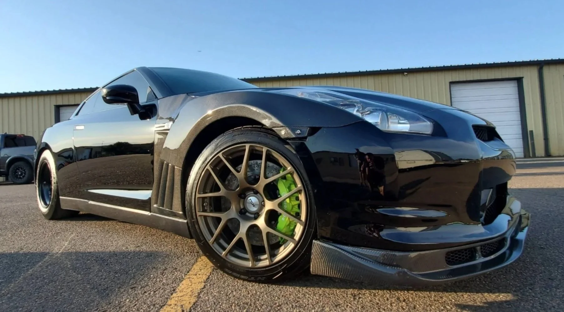 NISSAN GTR Black sports car with gold wheels and green brake calipers parked on an asphalt lot near a yellow warehouse.