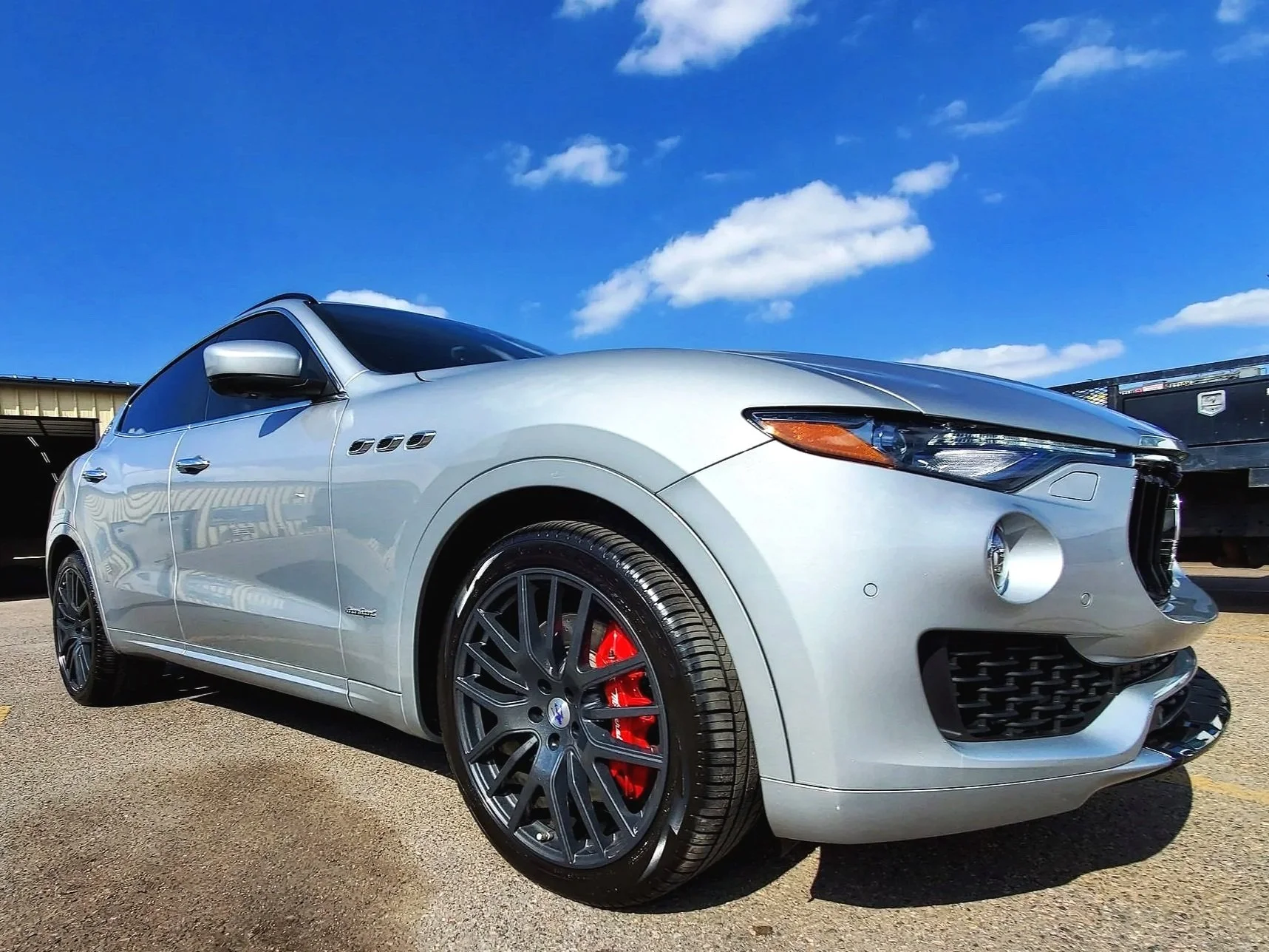 A silver luxury sedan MASERATI parked outdoors on a sunny day with a blue sky and scattered clouds. The car has black wheels, red brake calipers, and sleek design features.