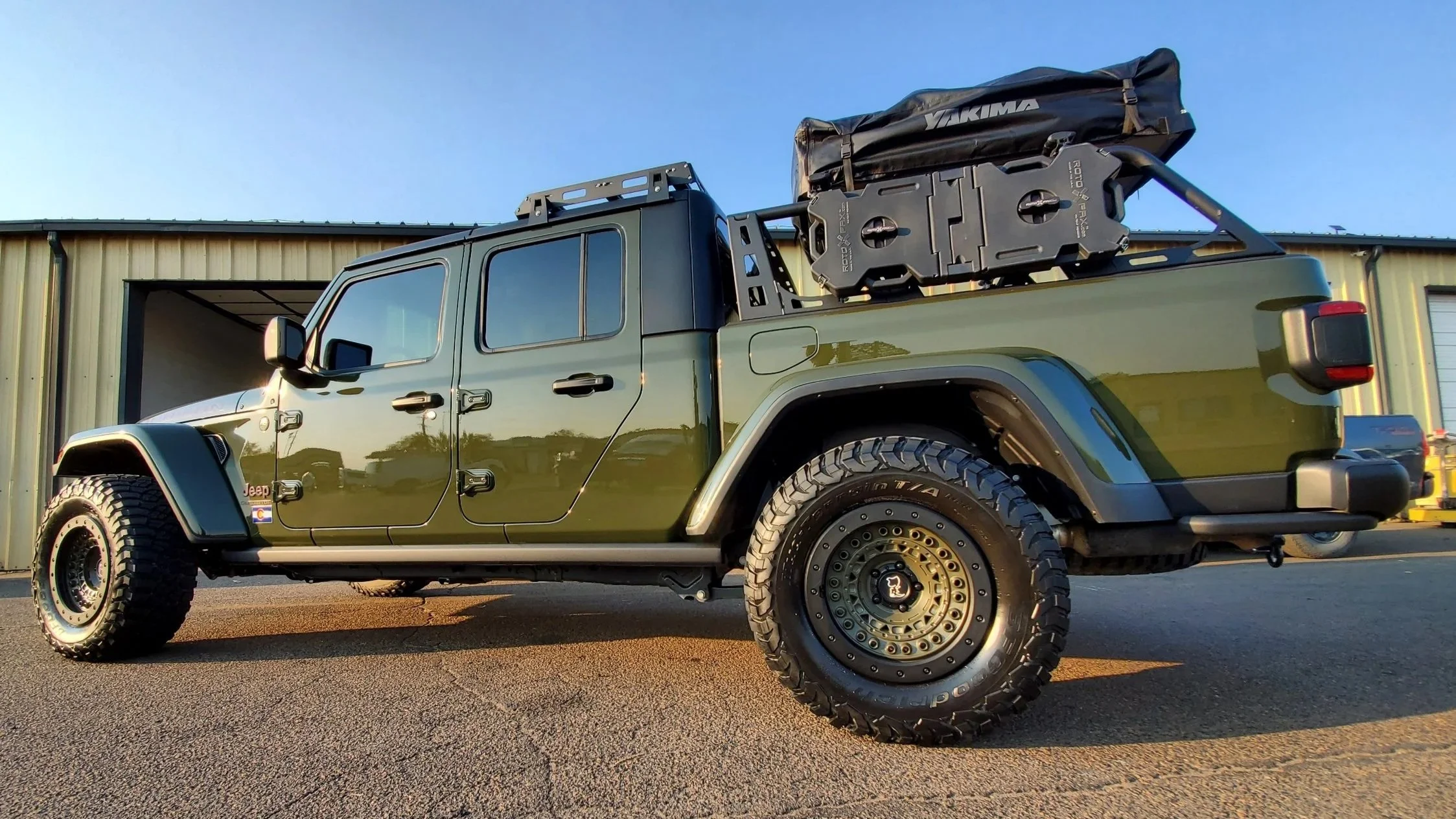 Green Jeep Gladiator pickup truck with off-road tires parked outside a metal building, carrying gear on a roof rack.