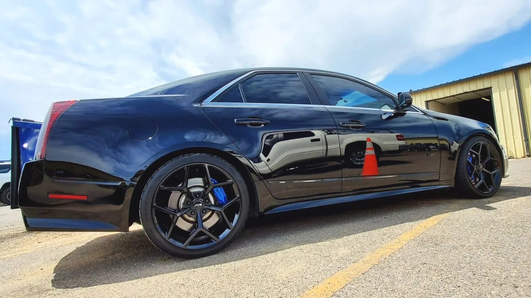A sleek black CTSV vehicle parked in a lot near a beige building, with a safety cone beside it and a partly cloudy sky overhead.